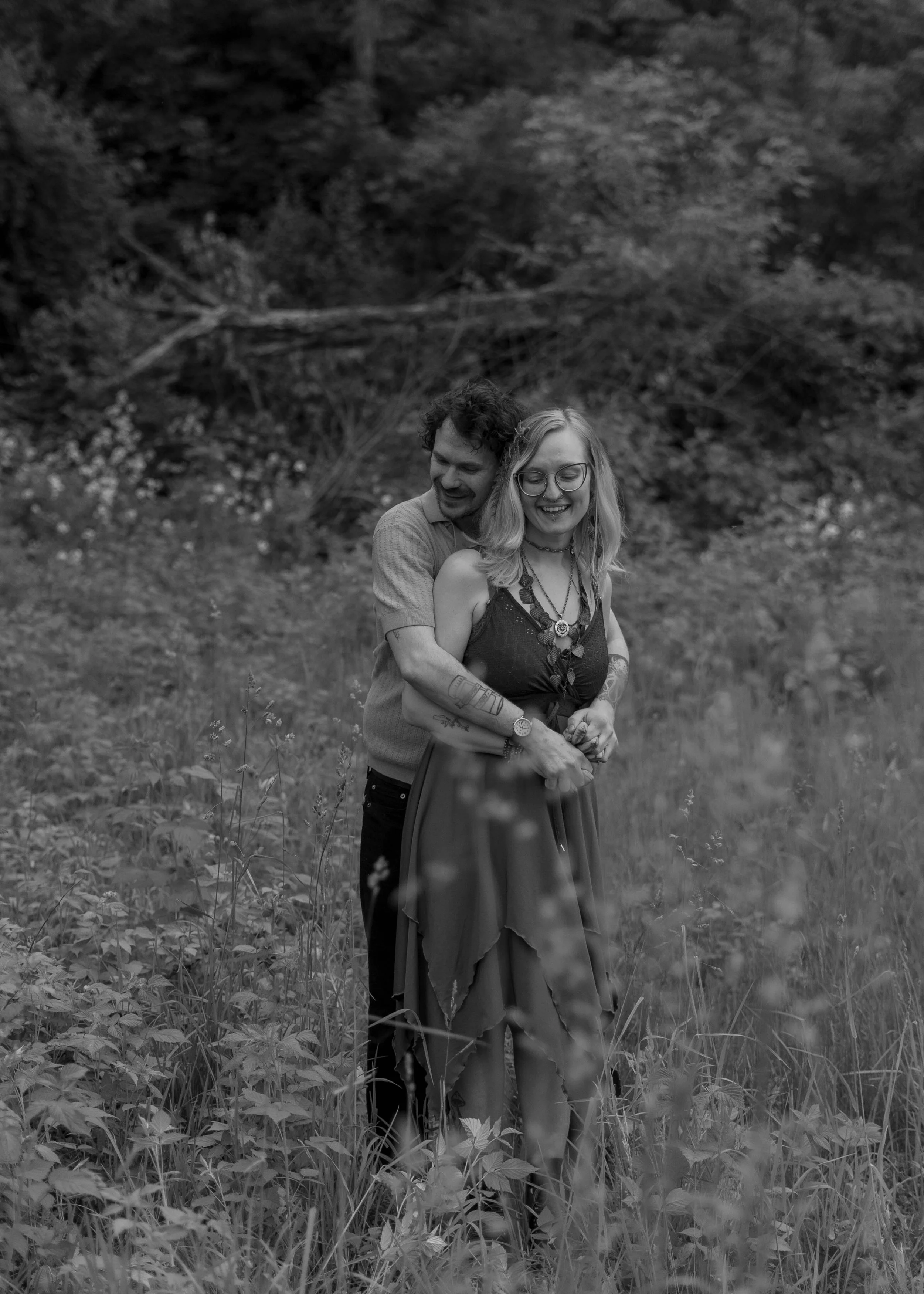A black-and-white photo of a man hugging a woman from behind in a grassy outdoor setting with trees and fallen branches in the background. Both are smiling.