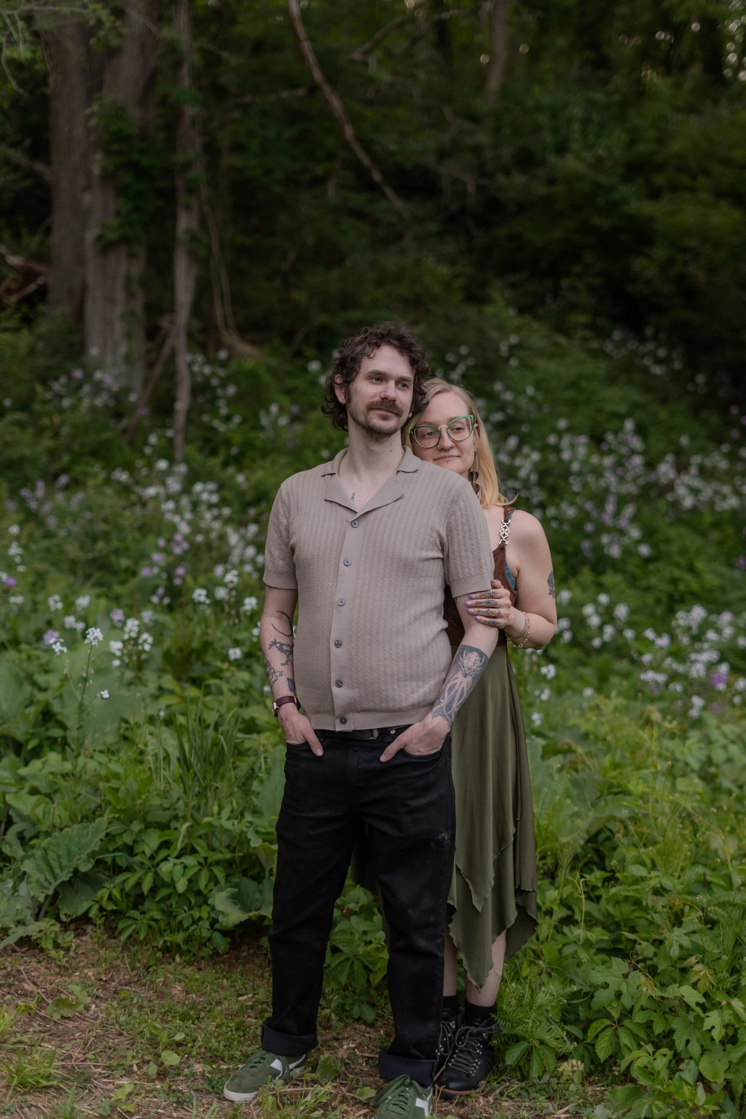 A couple standing in a lush, green outdoor setting with trees and flowers, embracing each other.
