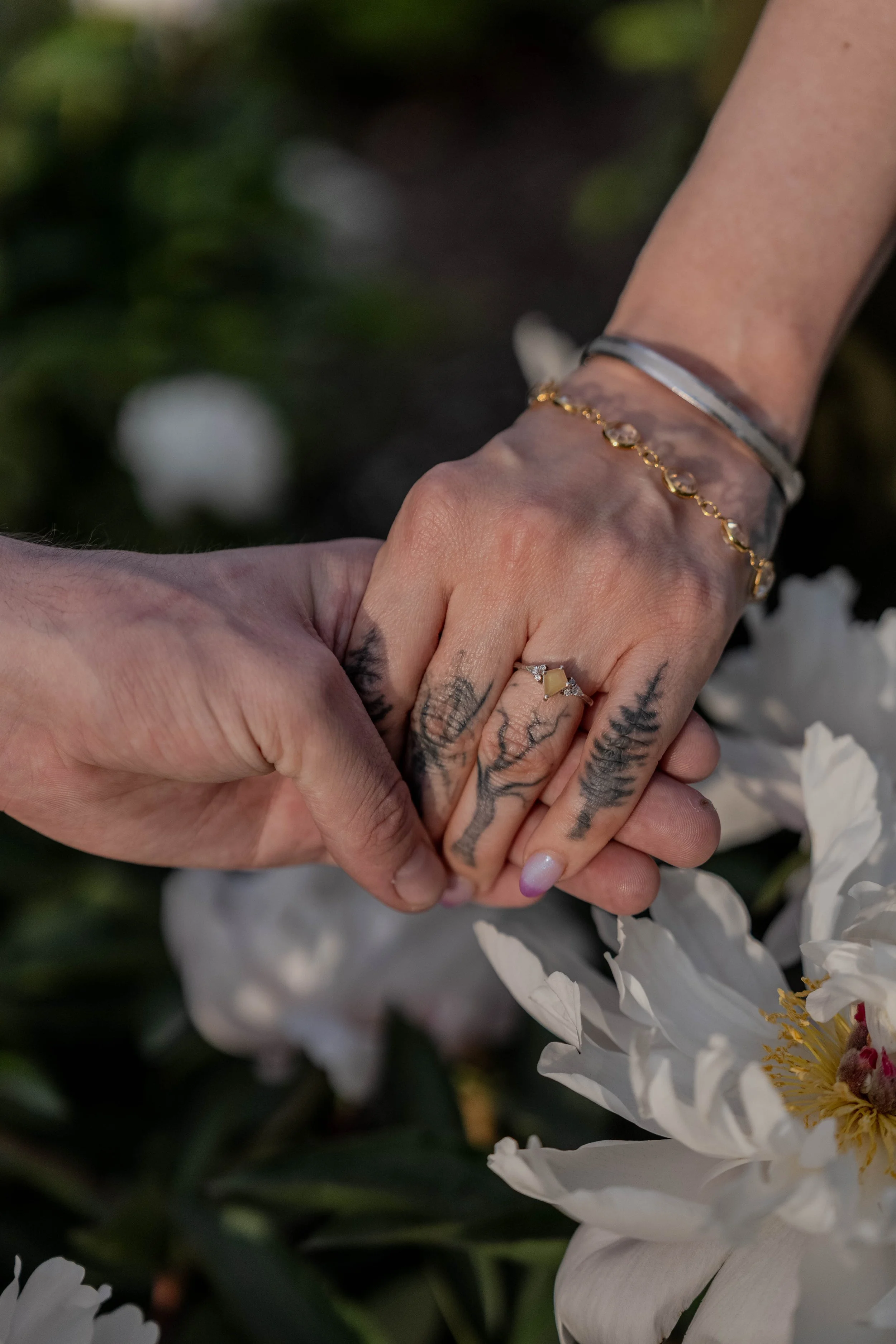 Two hands holding each other, one hand with tattoos and wearing rings, the other with jewelry, near white flowers with yellow centers.