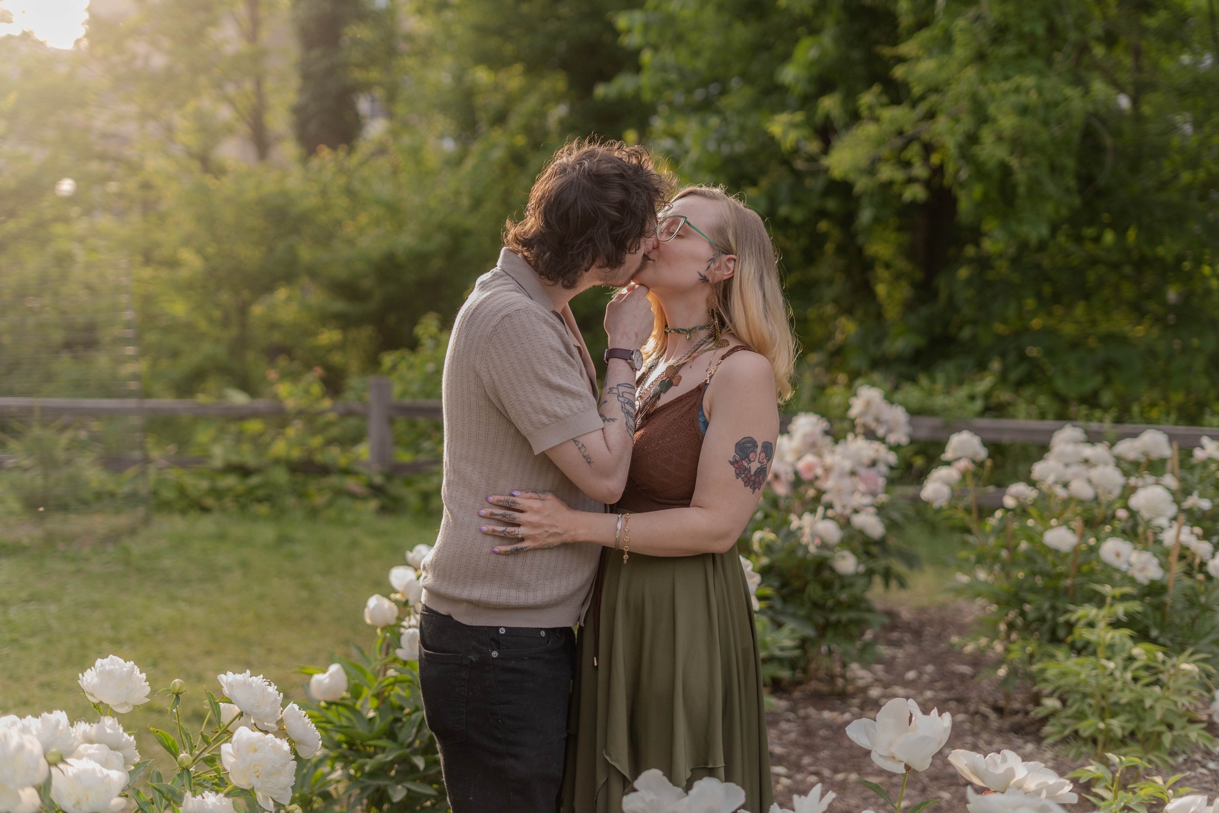 A couple sharing a kiss in a garden with white flowers and green trees in the background.