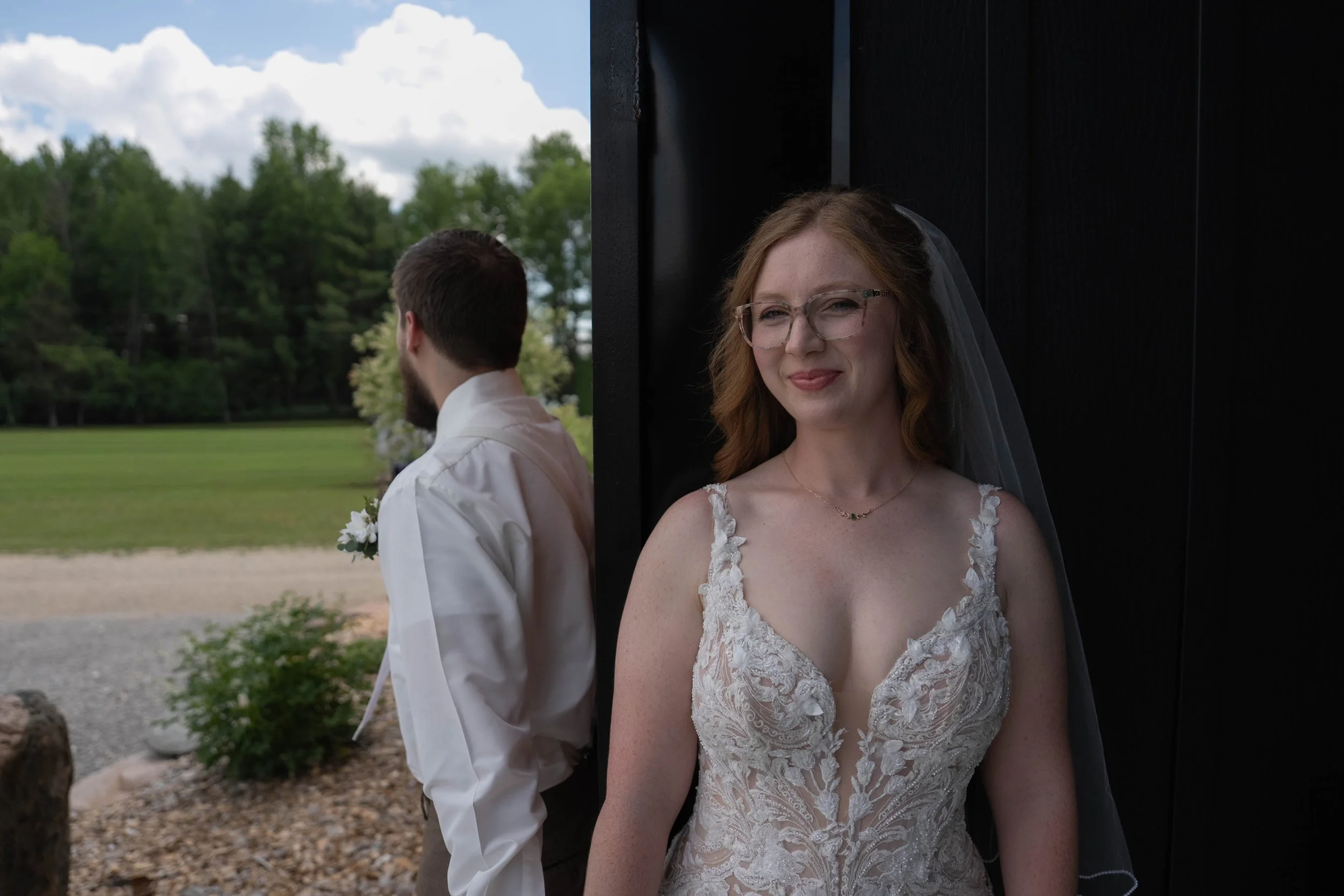 A bride with red hair and glasses, smiling with her eyes closed, stands beside a groom with his back to the camera, both outdoors in a park setting with trees and clouds in the background.