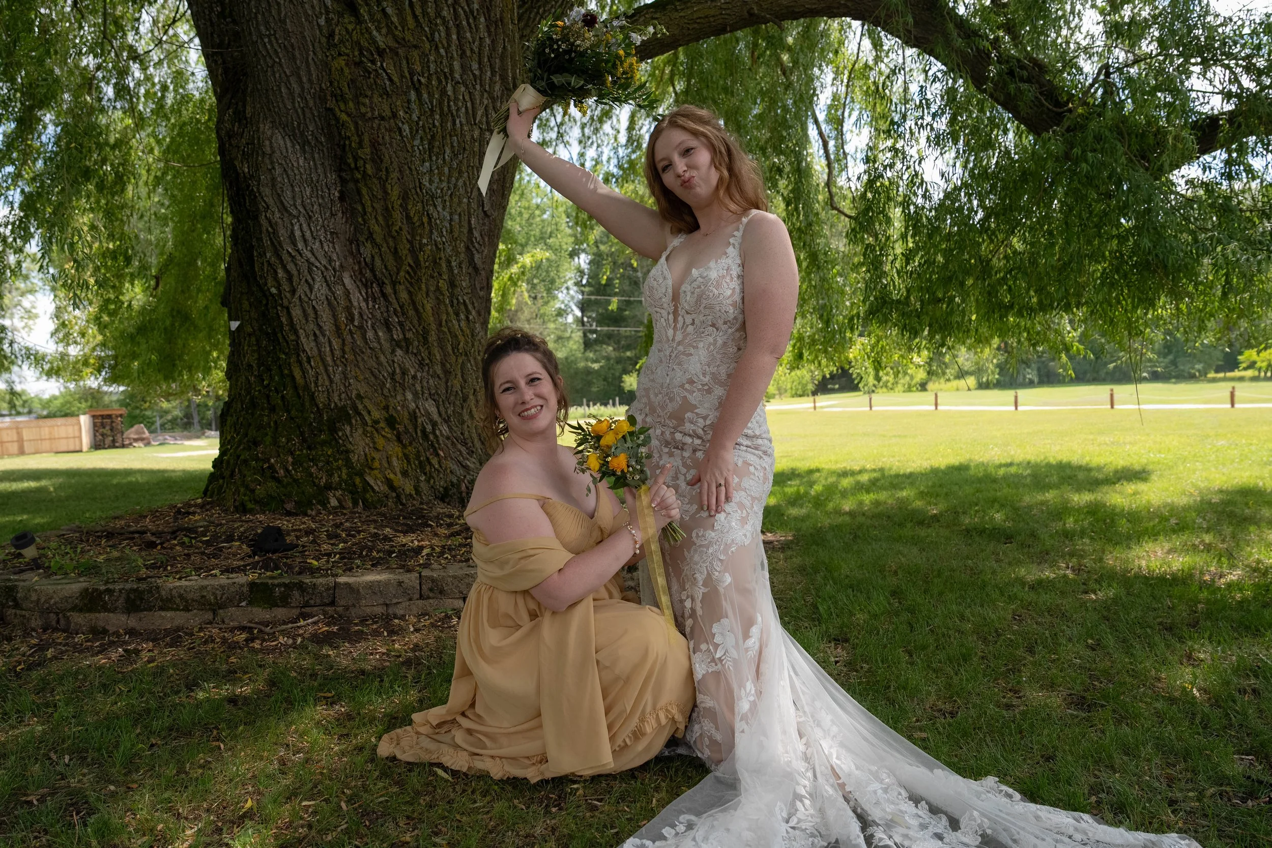 Two women, one in a wedding dress and the other in a gold gown, pose outdoors under a large tree. The woman in the wedding dress is standing and the other woman kneels holding a bouquet of yellow flowers. They are smiling and making playful facial ex