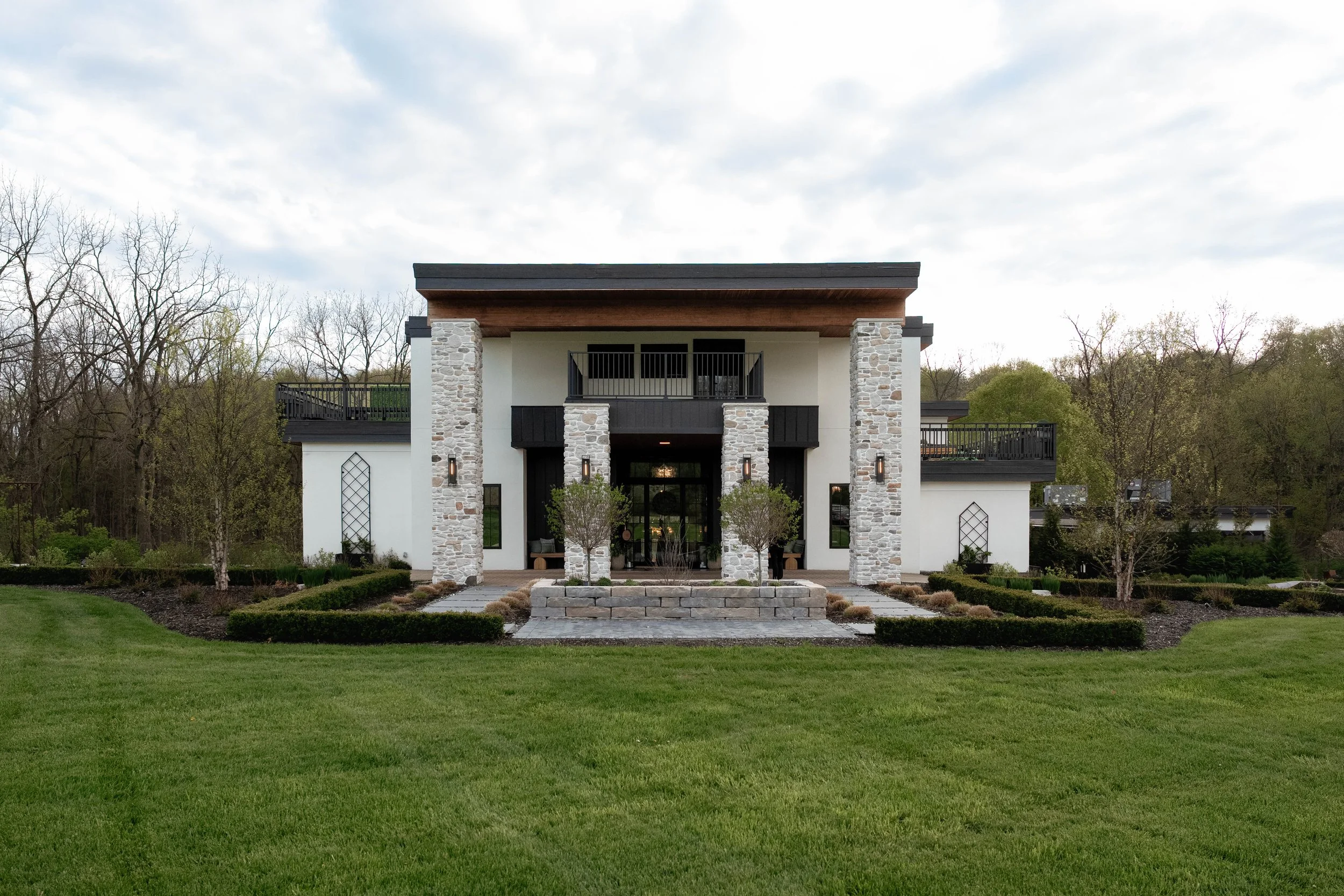 A modern two-story house with stone and white exterior, black accents, and a covered porch, surrounded by a green lawn and trees, under a cloudy sky.