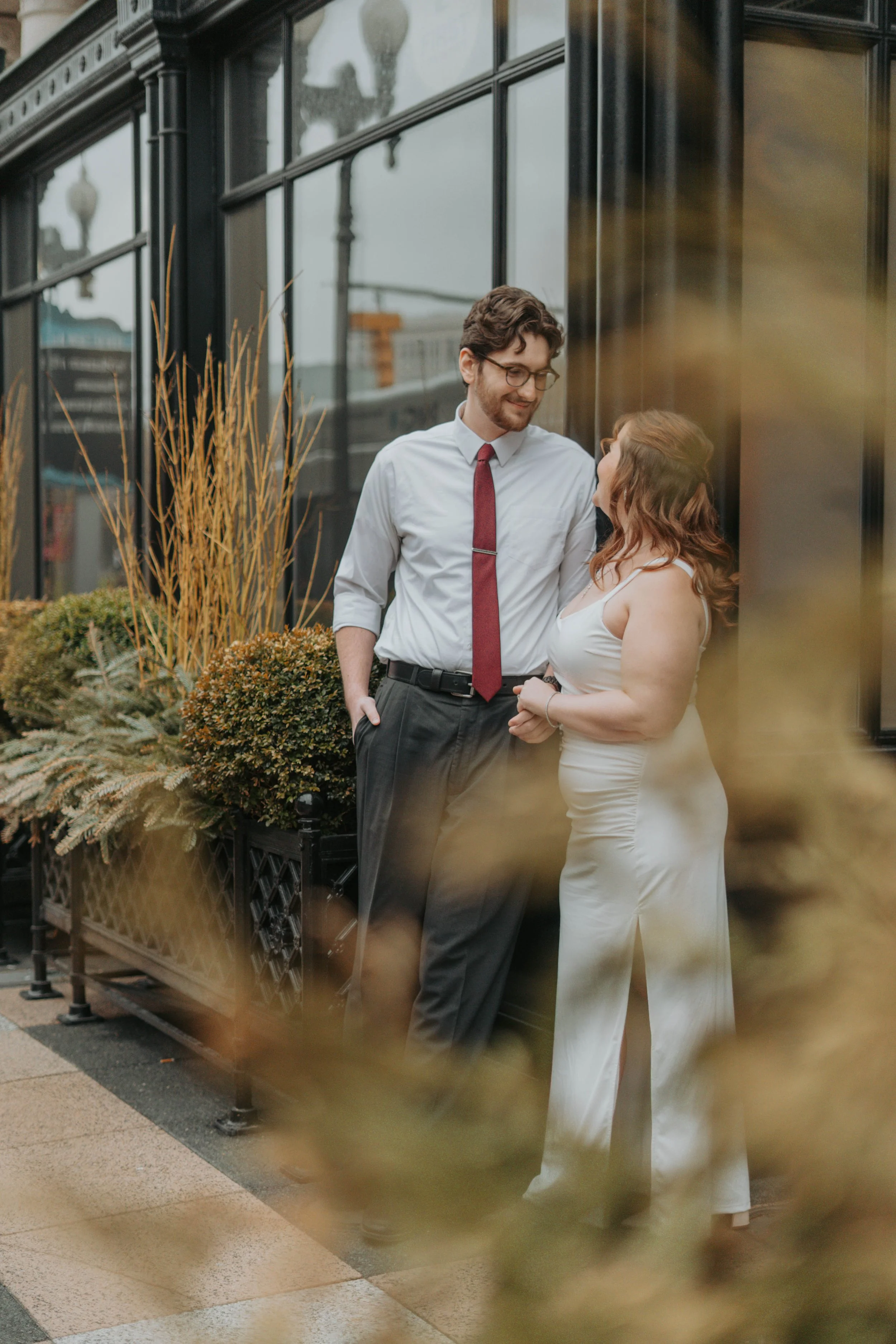 A man and woman are standing close together outside a building with large glass windows. The man is wearing glasses, a white shirt, and a red tie, while the woman is in a white dress. They are smiling and looking at each other, apparently engaged in 