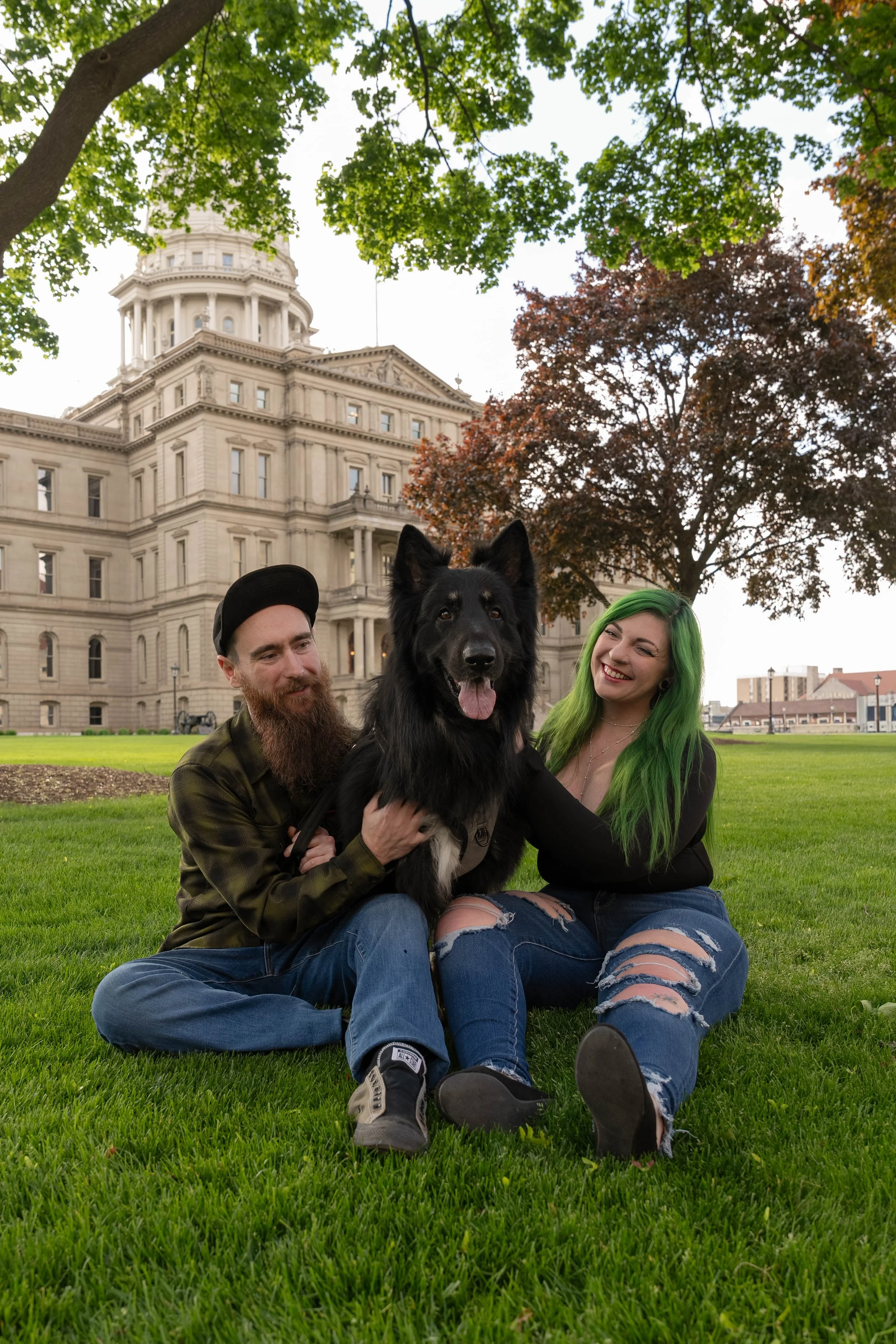 A couple and a large black dog sitting on green grass in front of a government building with a dome, surrounded by trees with green and reddish-brown leaves.