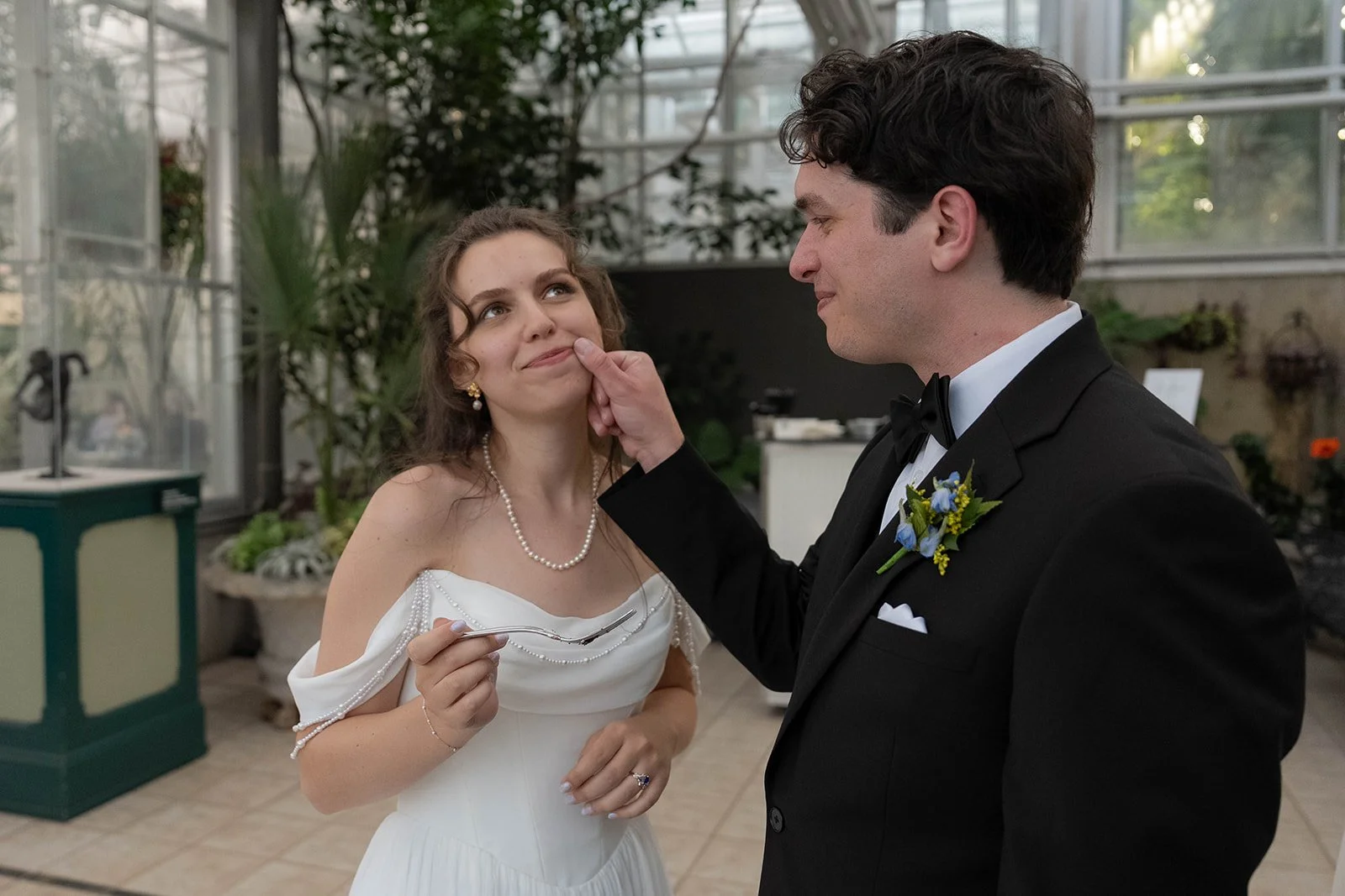A bride and groom in a greenhouse during their wedding. The groom is gently touching the bride's face. The bride is smiling and holding a fork, wearing a white dress with pearl accessories. The groom is dressed in a black tuxedo with a boutonnière.