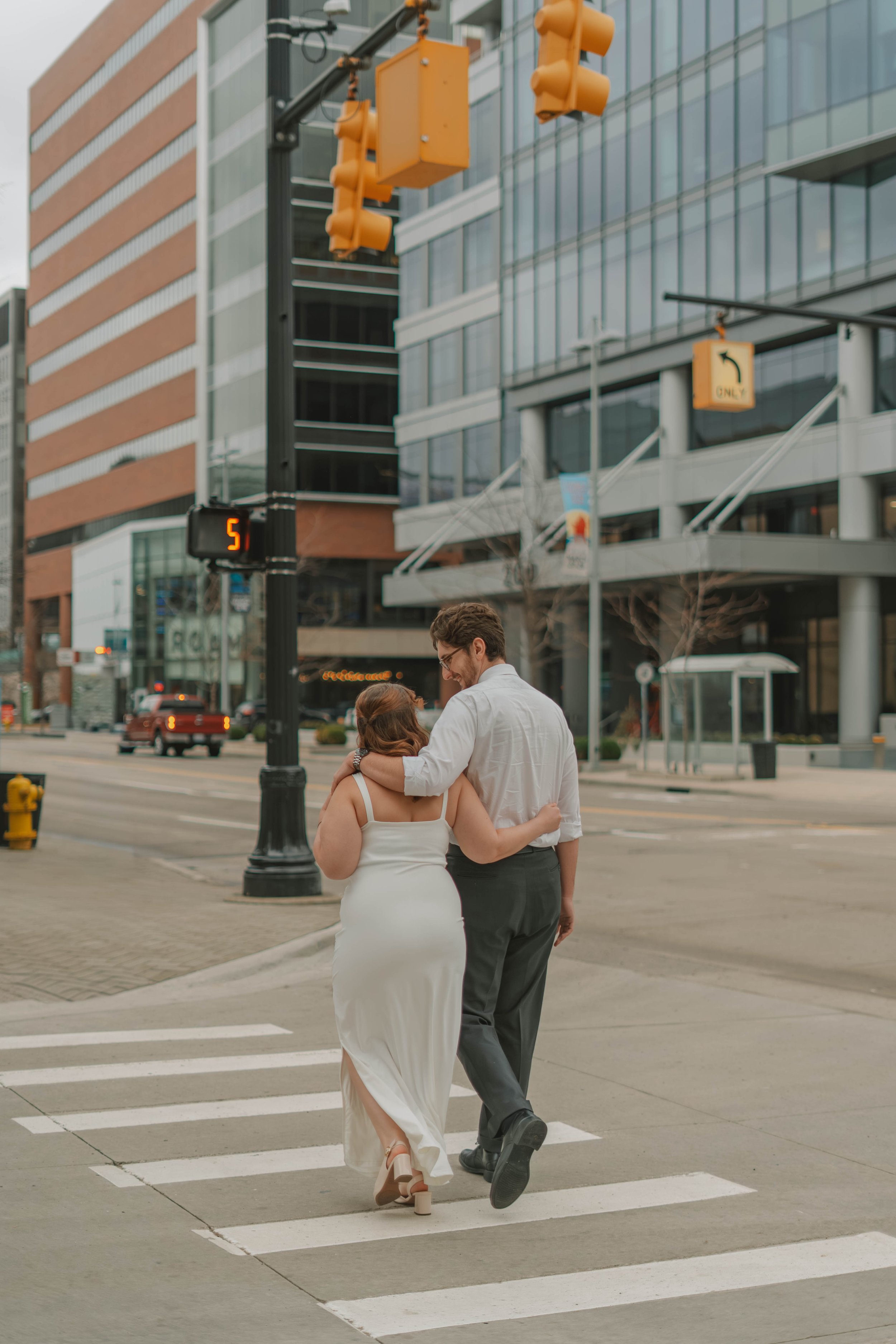 A couple dressed in wedding attire walking hand in hand across a city crosswalk. The woman is wearing a white dress with a slit and heels, and the man is wearing a white shirt and gray pants. They are smiling and embracing. The background features ta