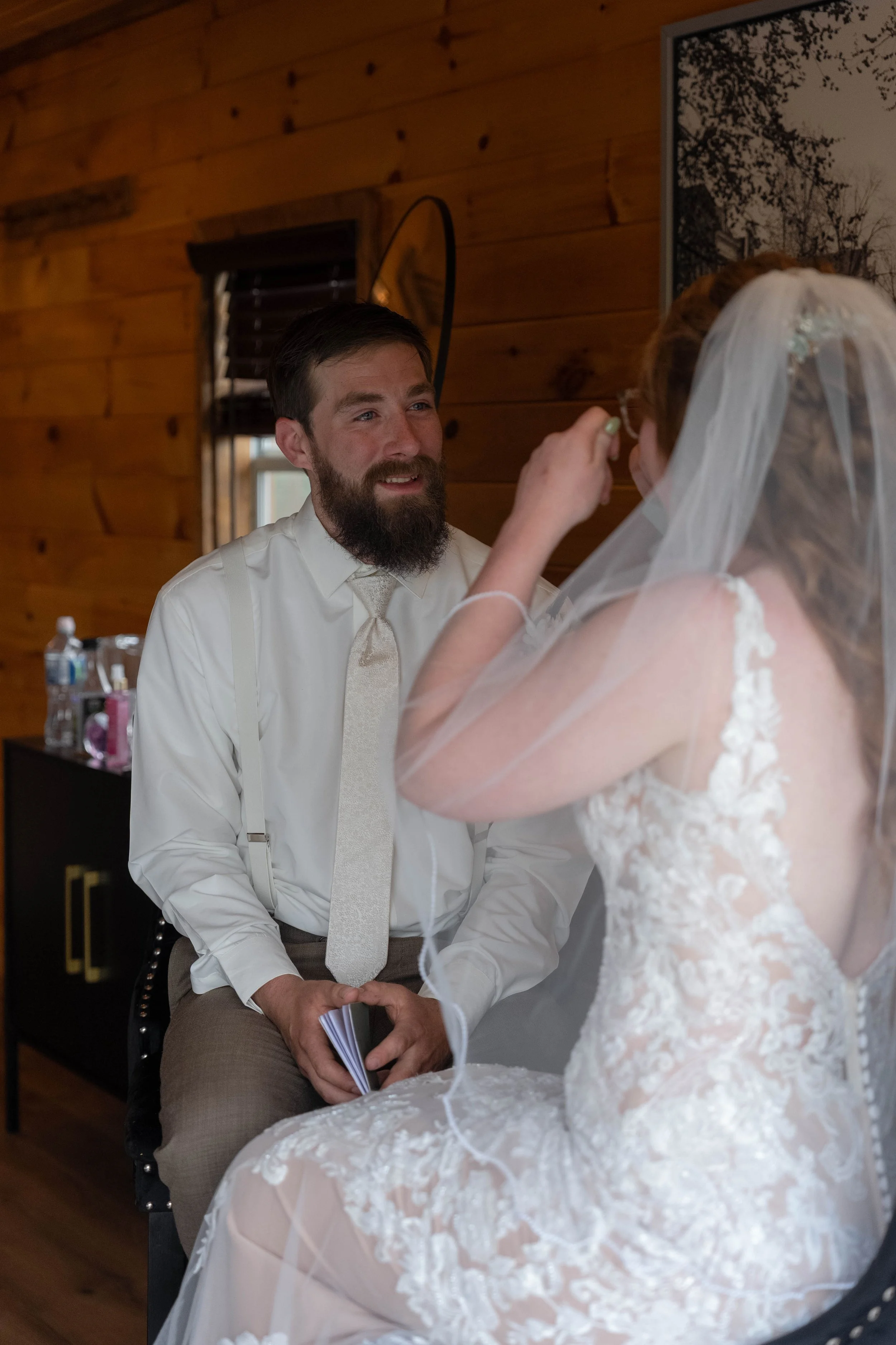 A groom with a beard and a white dress shirt smiles while sitting in front of a bride with a veil, in a wooden-paneled room during a wedding ceremony.