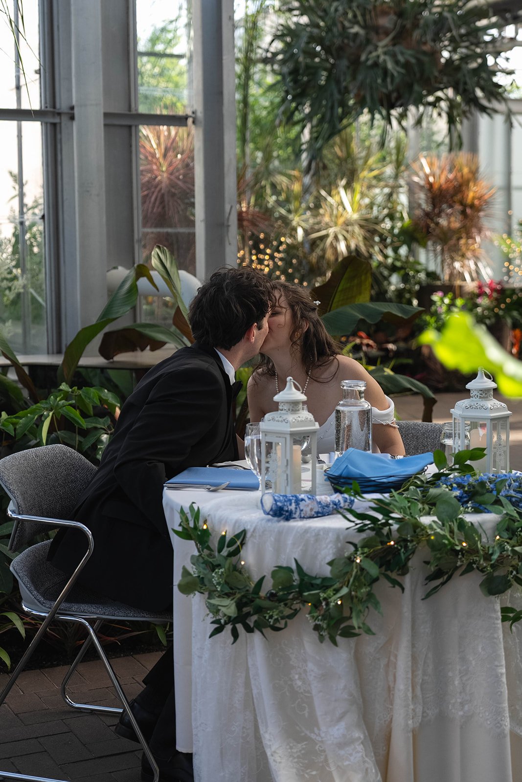 A couple in wedding attire sharing a kiss at a decorated table in a greenhouse or conservatory filled with plants and natural light.