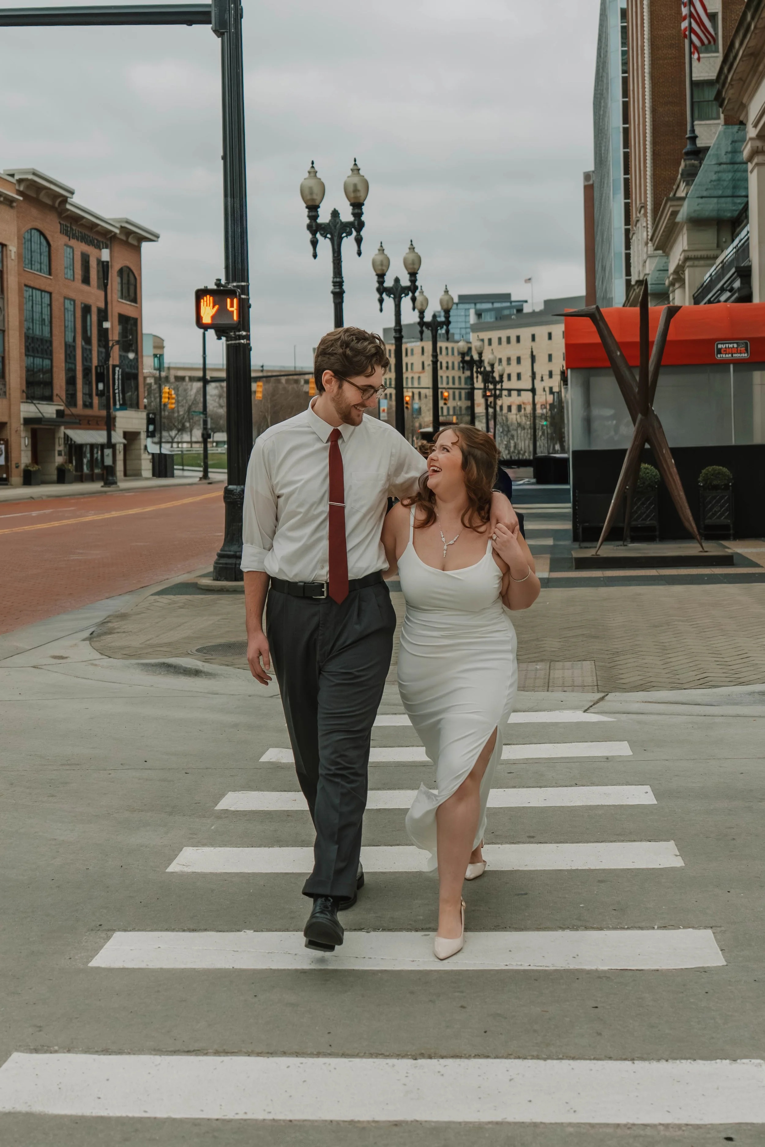 A couple is walking together across a crosswalk on a city street, smiling and looking at each other. The man is wearing glasses, a white shirt, and dark pants with a red tie. The woman is wearing a white dress with a slit and beige heels. The backgro