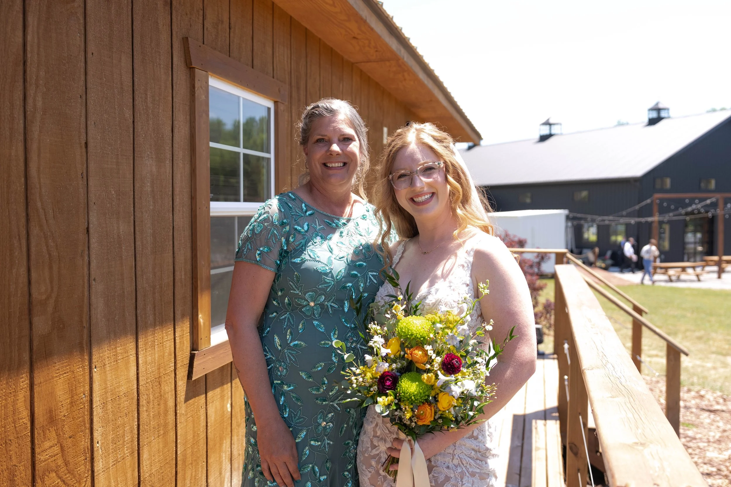 Two women smiling, one in a teal embroidered dress and the other in a white wedding gown holding a colorful bouquet, standing outside near a wooden building with a wooden railing.