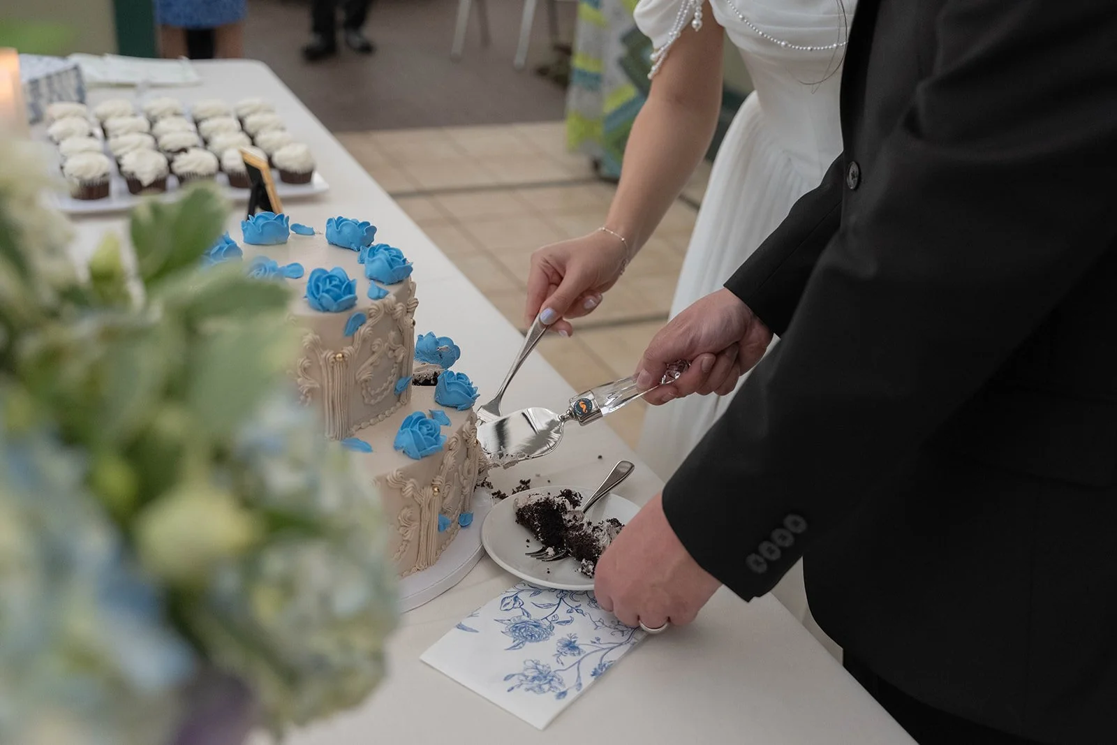 A couple is cutting a wedding cake decorated with blue roses during a wedding reception. Cupcakes with white frosting are visible in the background.