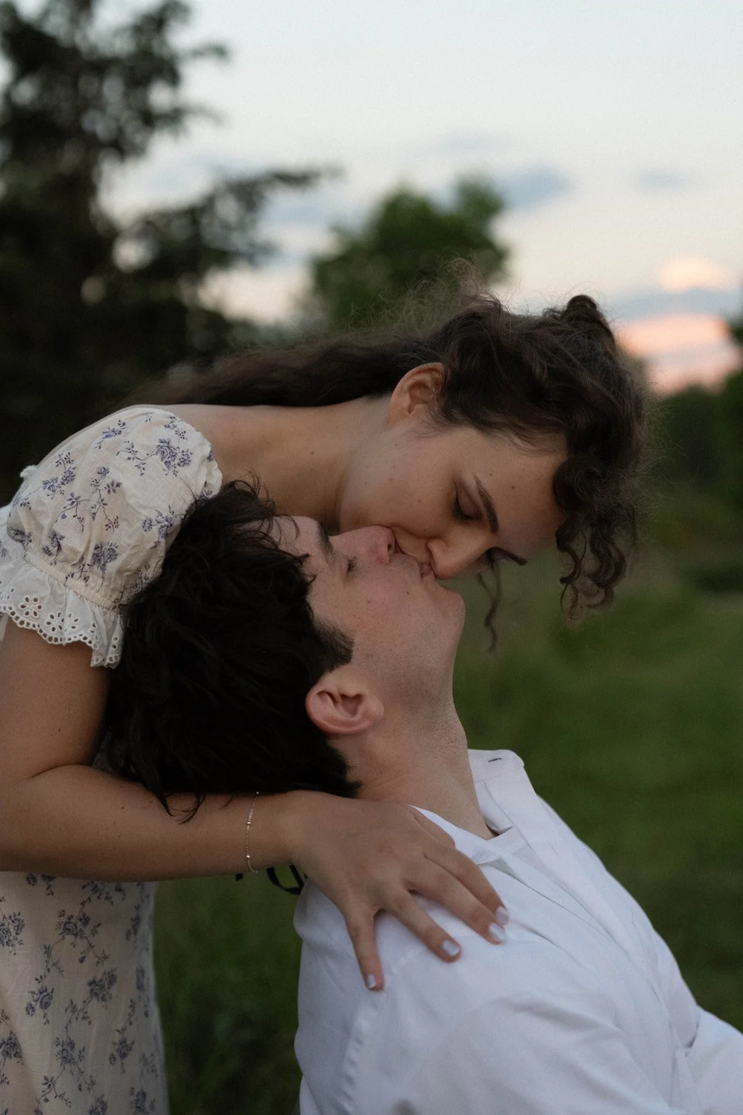 A couple sharing a kiss outdoors at sunset, with the woman leaning over the man and both smiling.