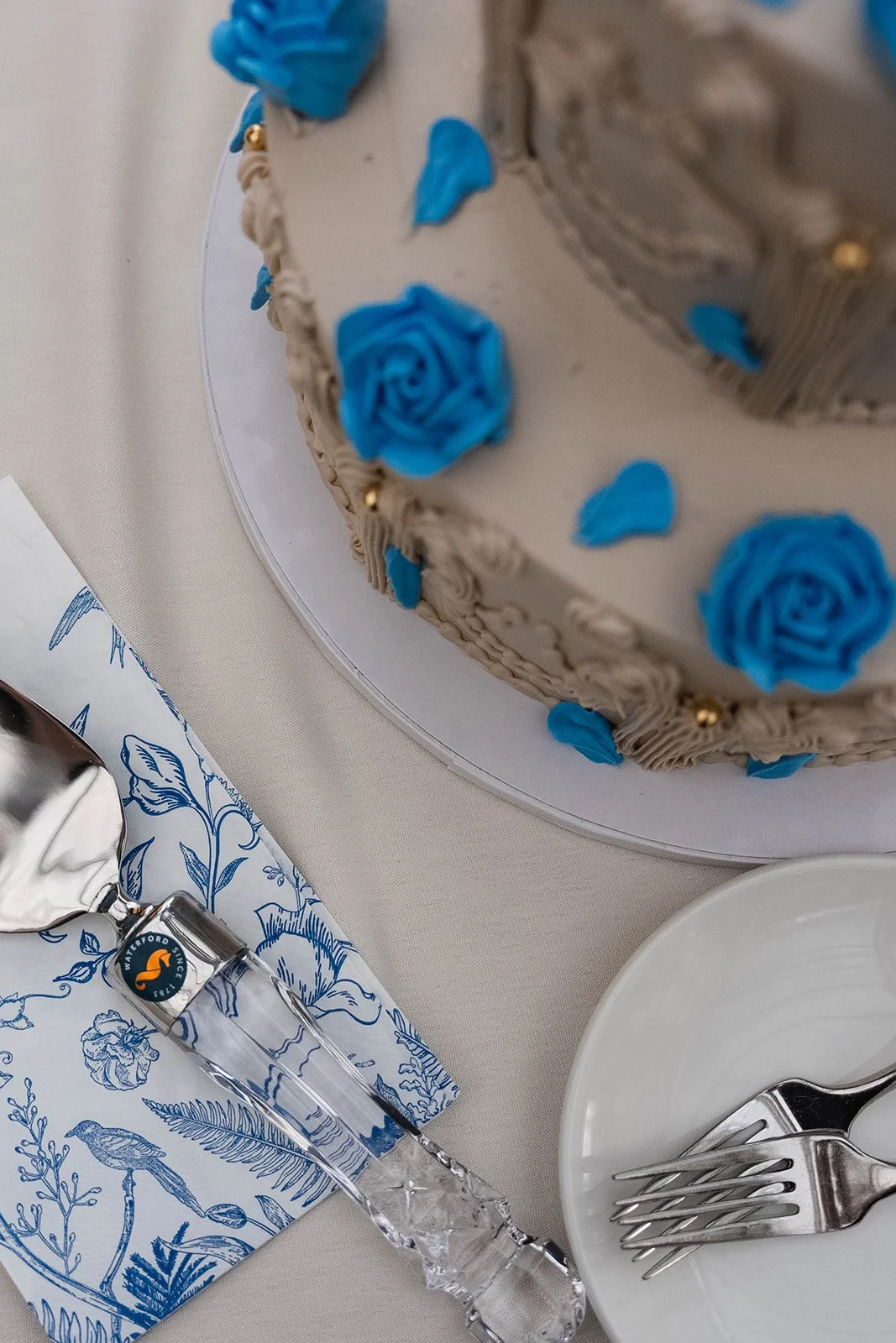 A close-up of a decorated cake with blue roses and beige piping, placed on a white cake stand. Part of a table setting with a blue and white floral napkin, a crystal water glass, and a white plate with a fork and spoon