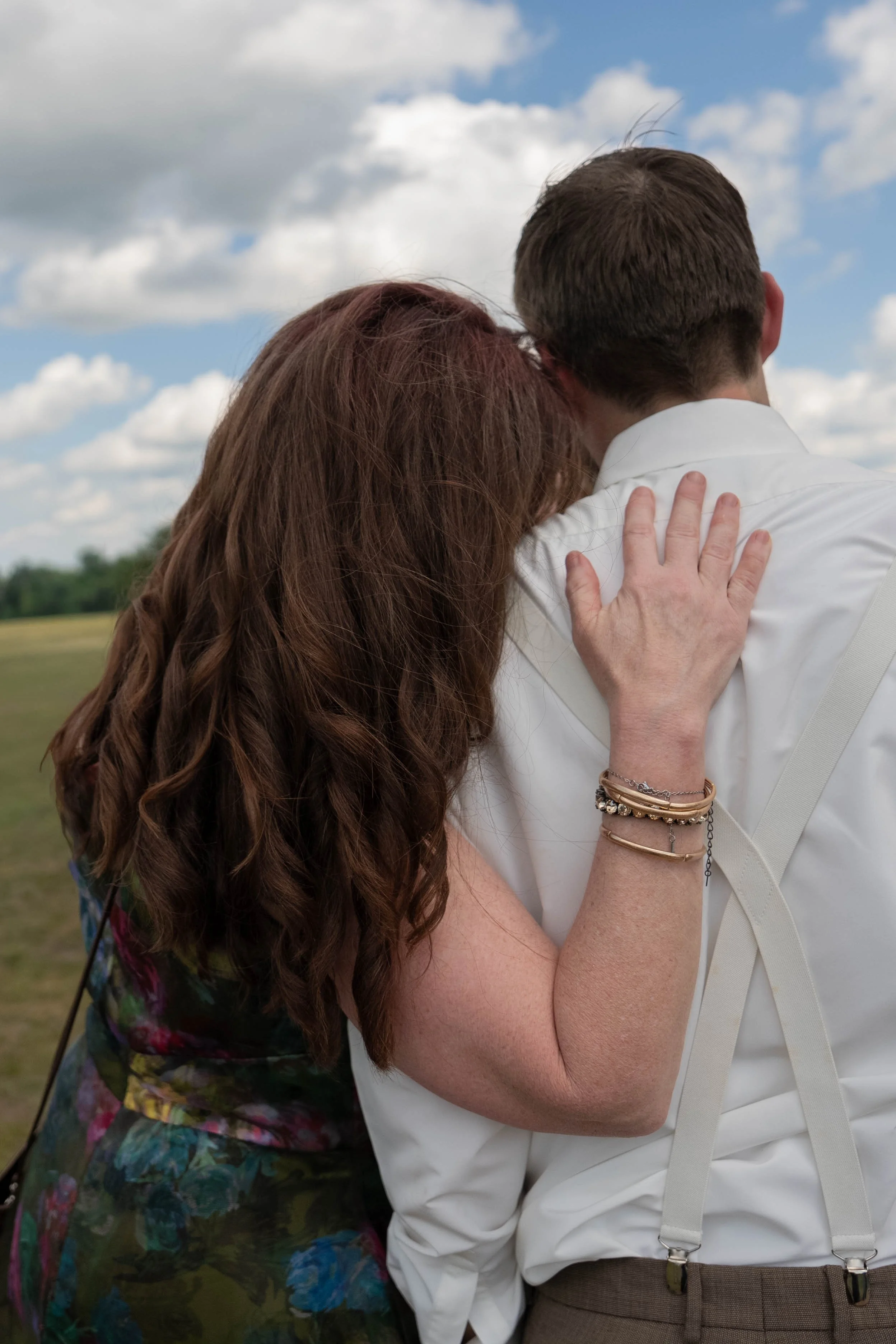 A couple embracing outdoors, with the woman resting her head on the man's shoulder. The man is wearing a white shirt with suspenders, and the woman has long, curly reddish-brown hair. The background features a cloudy sky and open field.