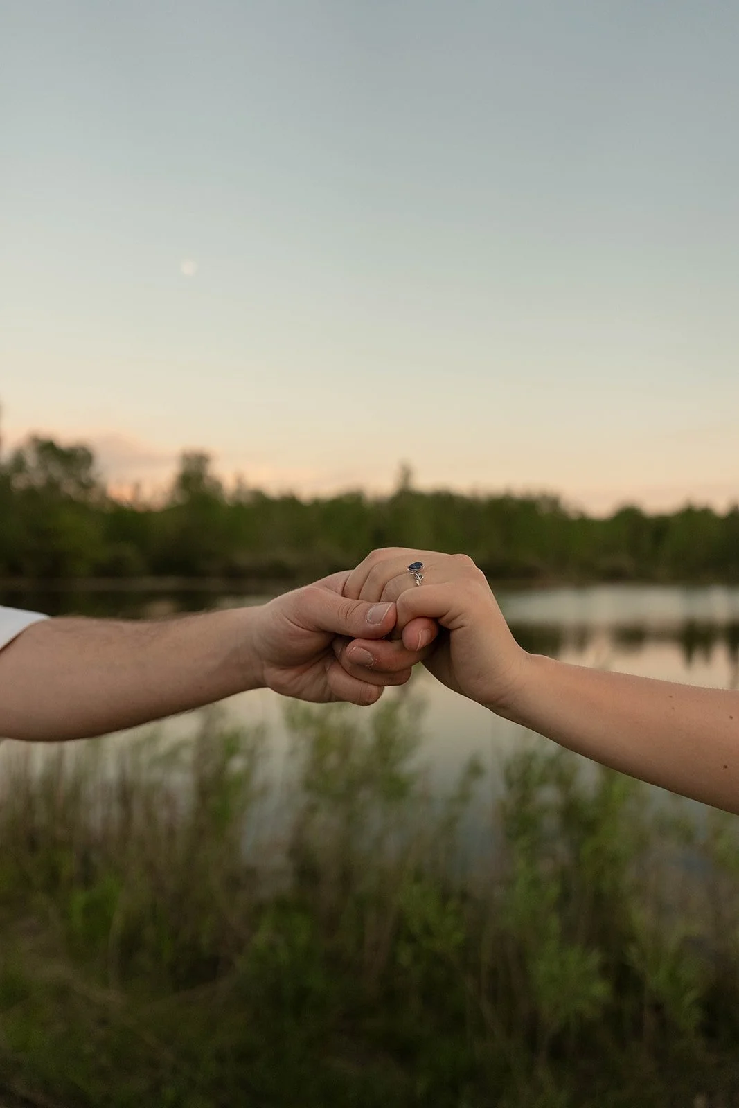Two people holding hands with a ring, standing outdoors near water with trees, during sunset or dusk.