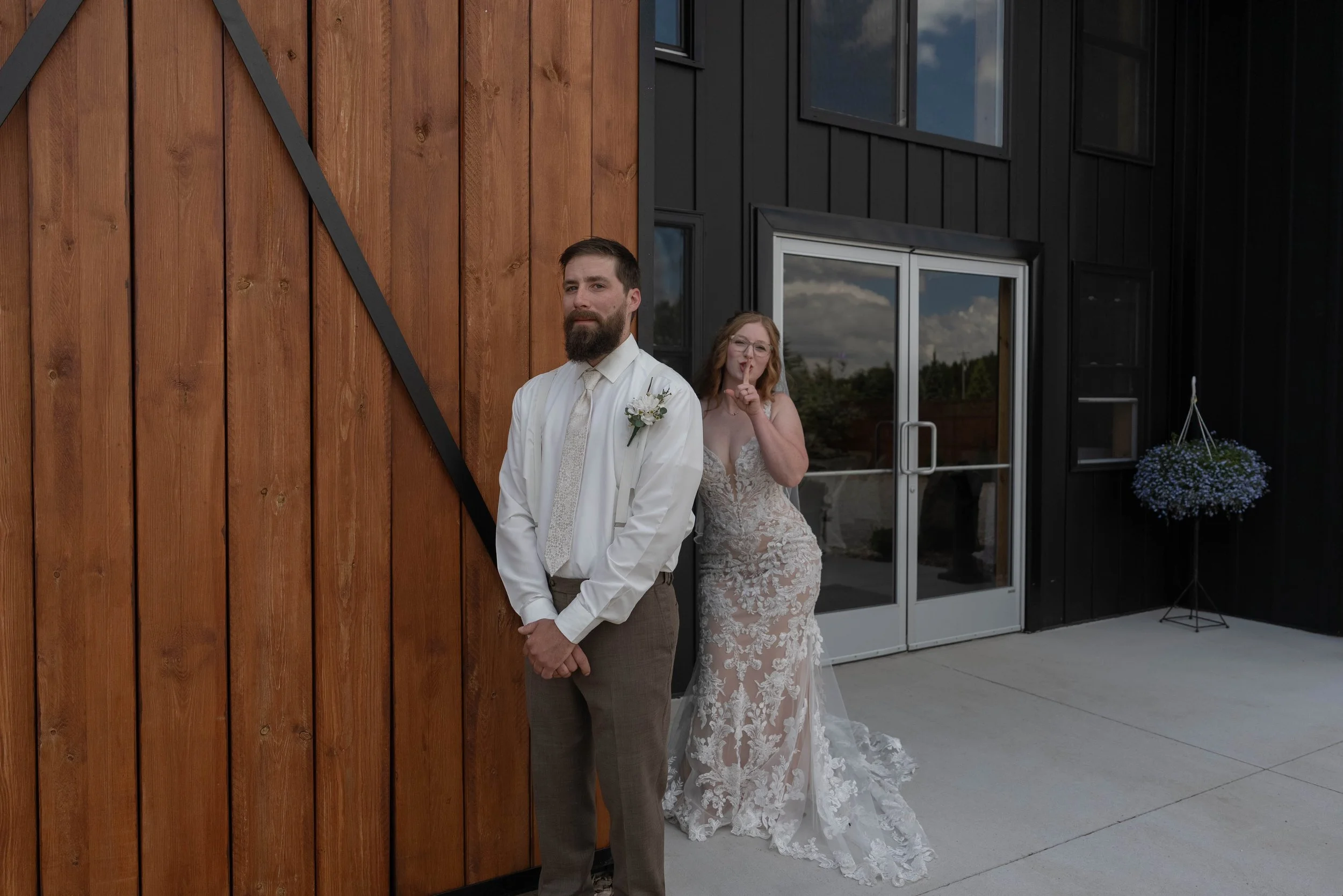 A man with a beard in a white shirt and tie stands in front of a wooden wall, while a woman in a wedding dress with lace details makes a shushing gesture behind him outside a modern building with black siding and glass doors.