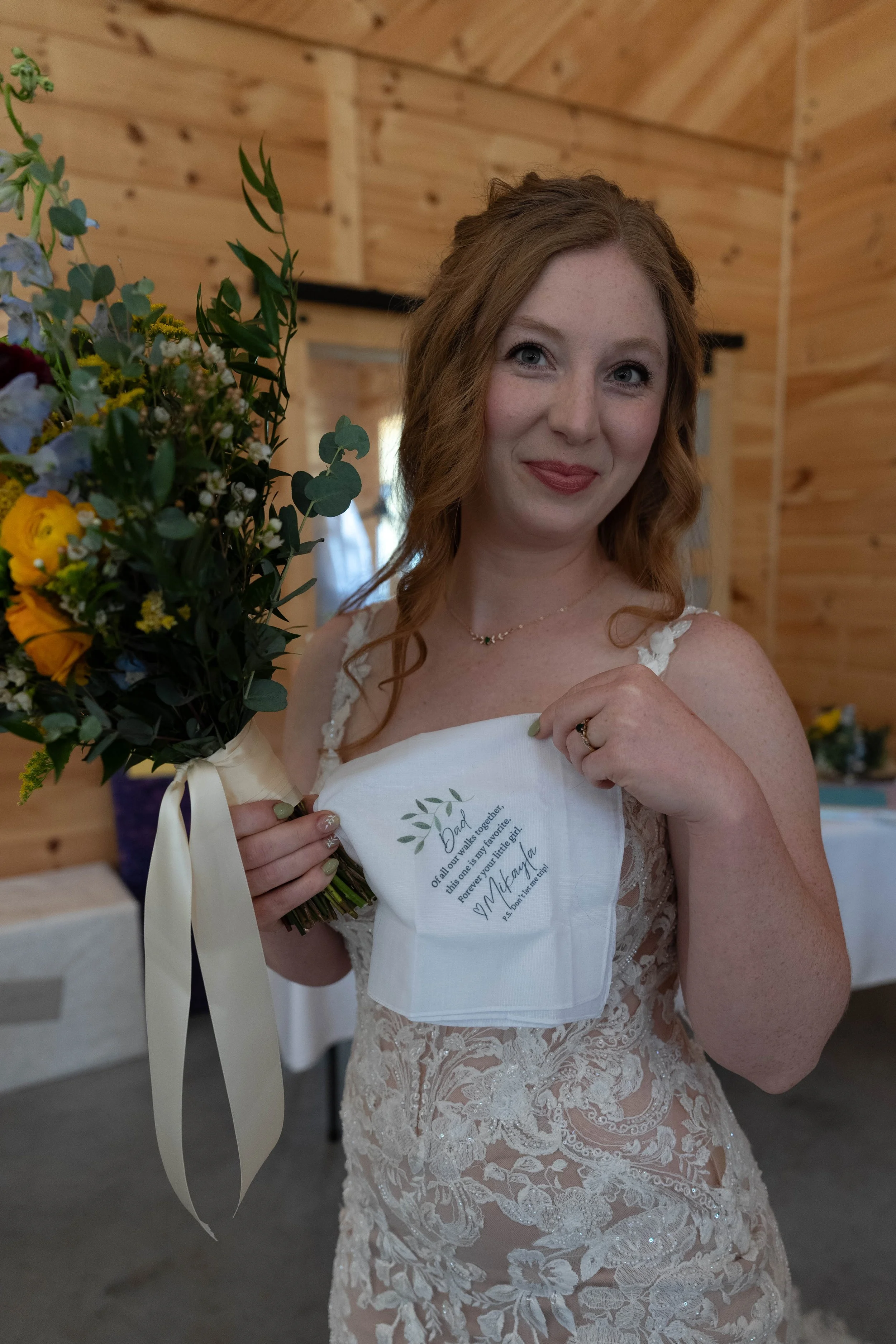 A woman in a wedding dress holding a bouquet of flowers and a personalized handkerchief with a heartfelt message inside a wooden building.
