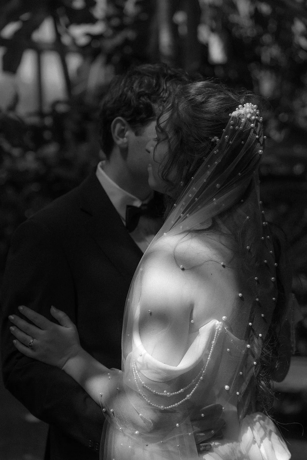 A black and white photo of a bride and groom embracing during their wedding, with the groom wearing a tuxedo and the bride wearing a wedding dress with a sheer veil decorated with pearls.