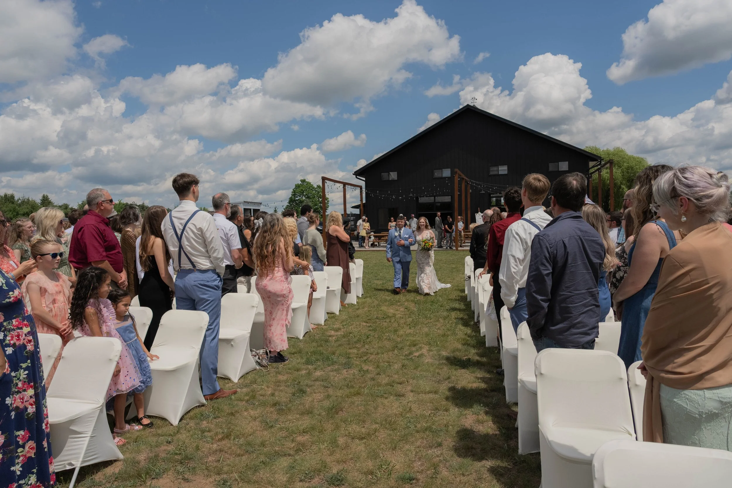 A group of people gathered outdoors for a wedding ceremony on a sunny day, with a black barn and string lights in the background.