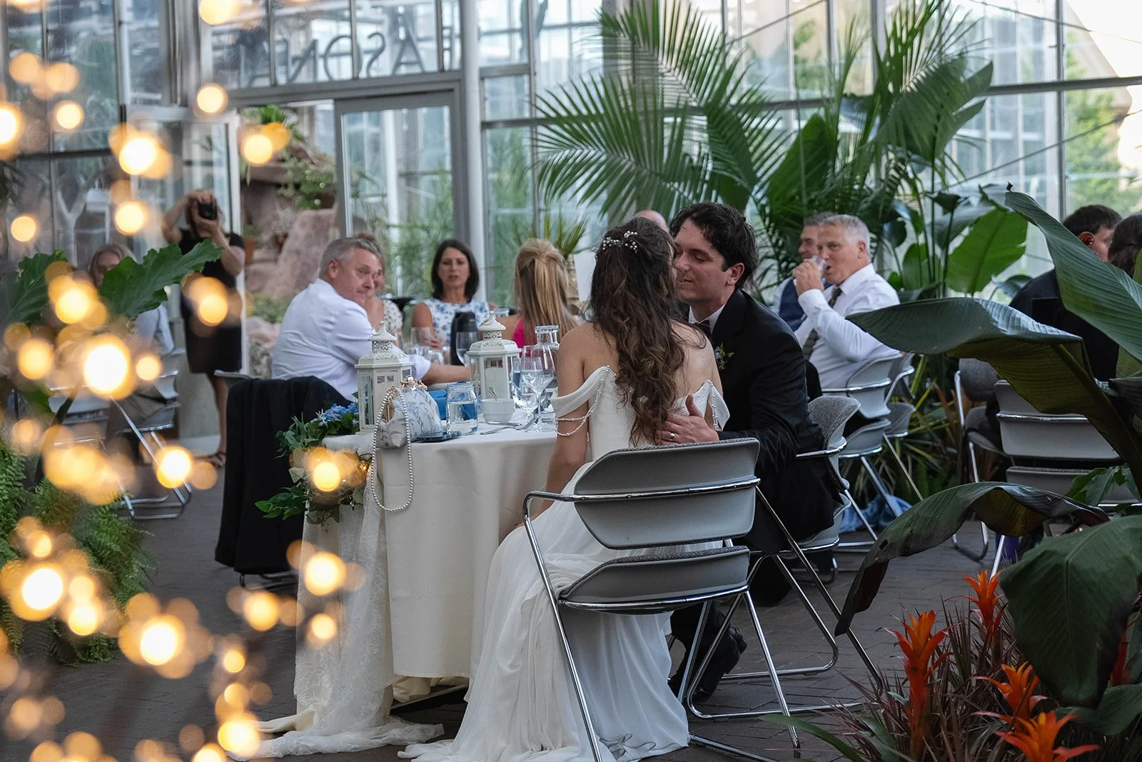 A bride and groom sitting at a table during their wedding reception, surrounded by guests, with large green plants and glass walls in a bright conservatory-style venue.