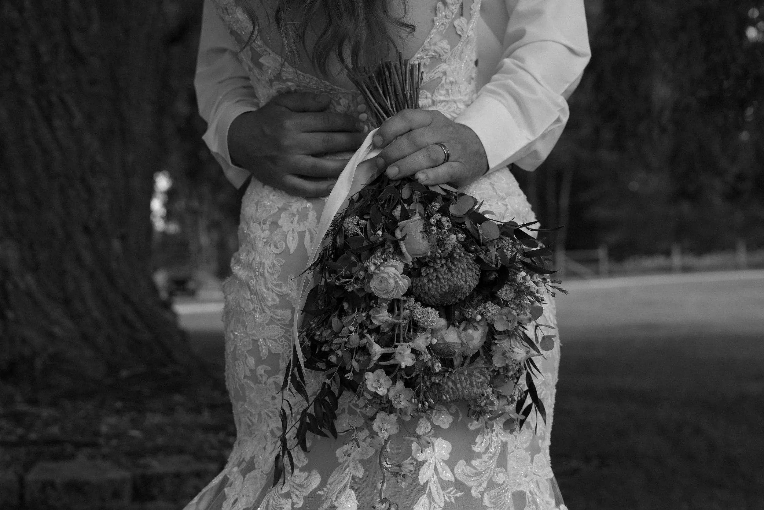 A bride in a lace dress holding a bouquet of flowers, with hands gently clasped around the stems, hidden face, outdoor setting with trees visible in the background.