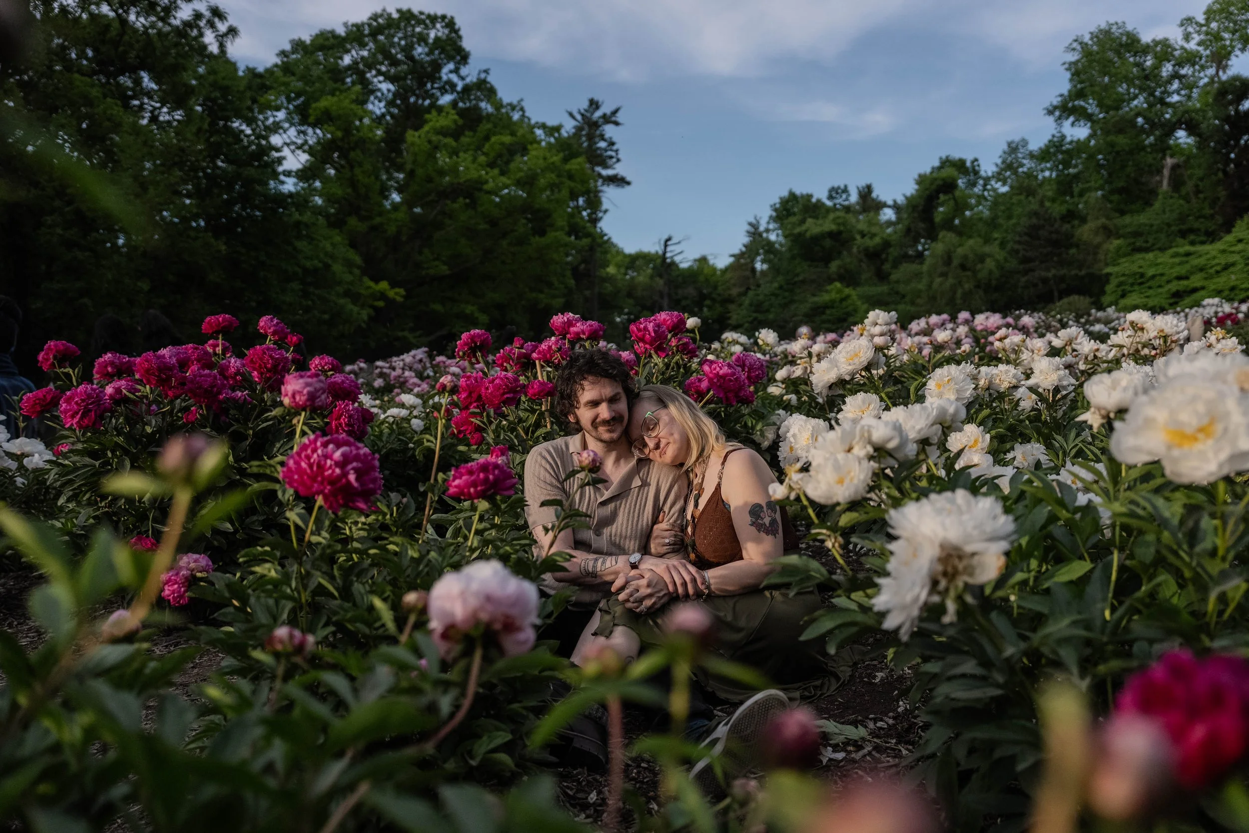 A couple sitting amidst pink and white peony flowers in a garden with green trees and a partly cloudy sky in the background.