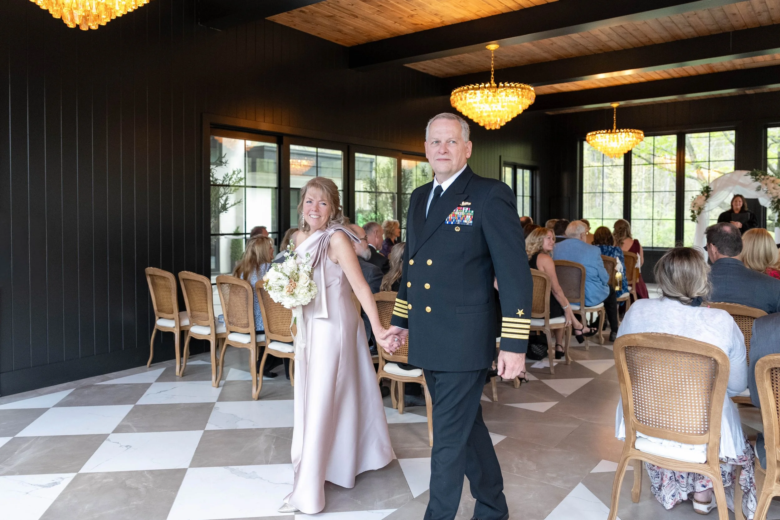 A couple holding hands at a wedding ceremony, with guests seated in the background inside a venue with large windows and hanging chandeliers.