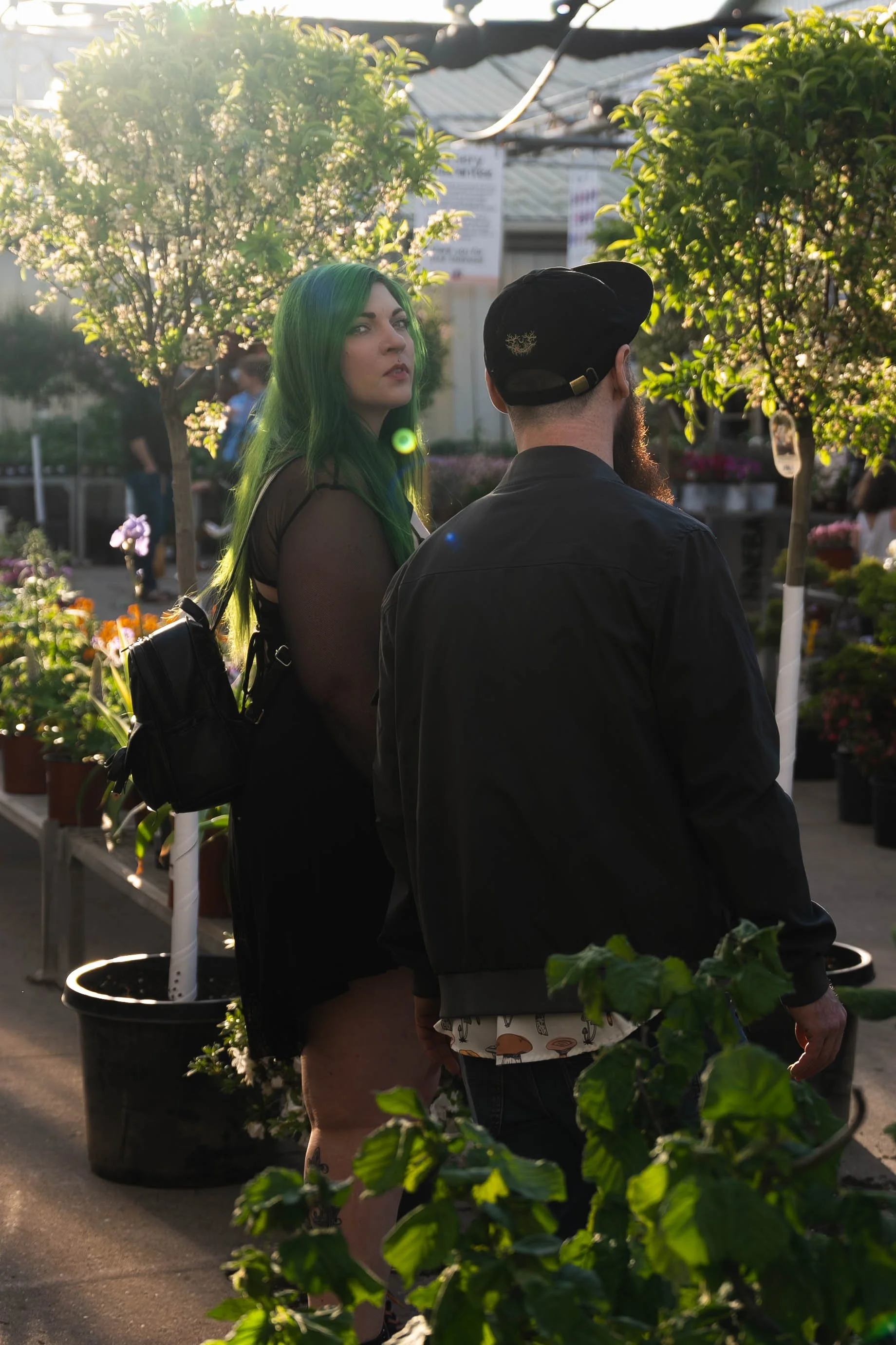 A woman with green hair and a man with a beard and cap standing outdoors in a garden center, surrounded by trees and plants, with sunlight streaming through.
