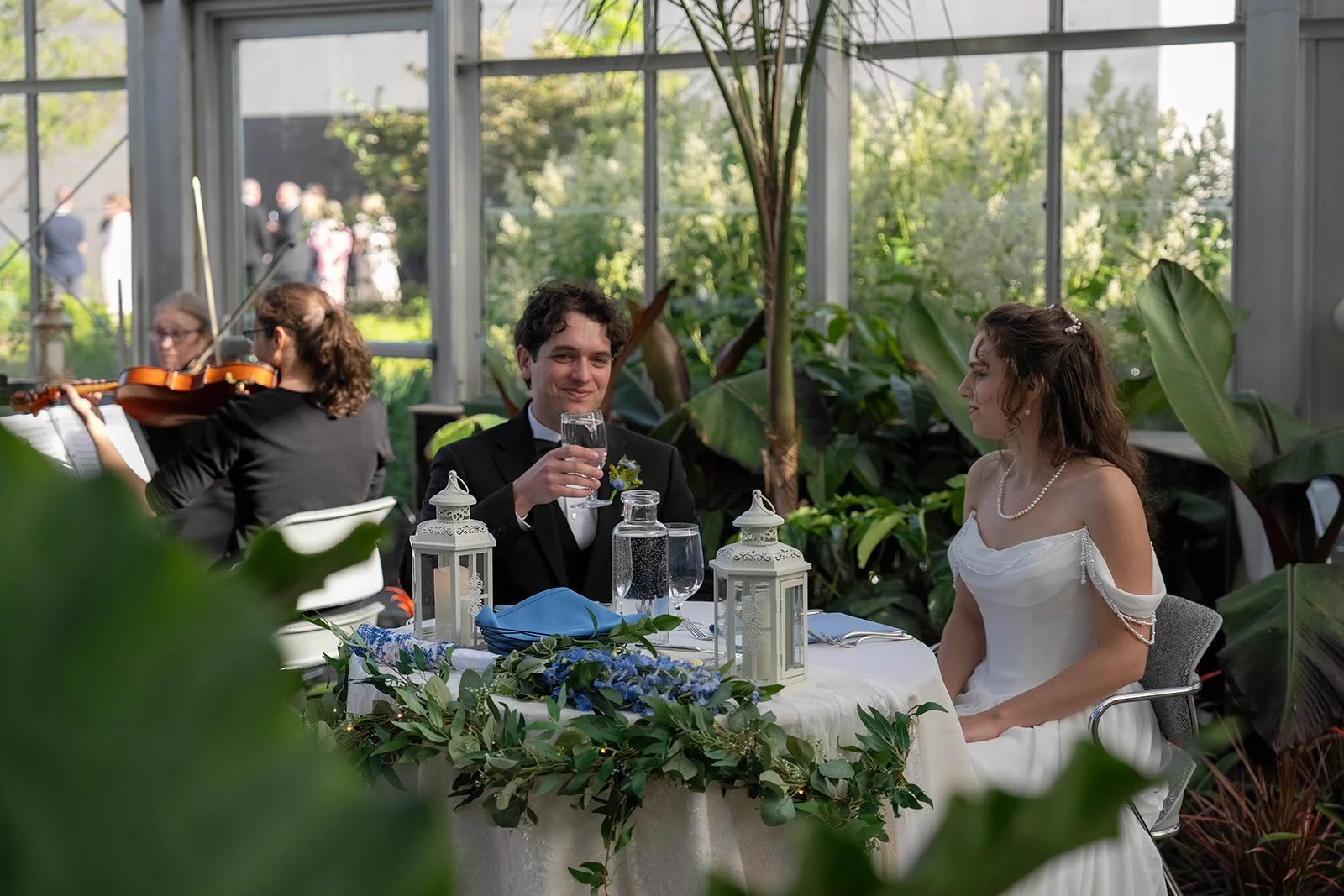 A bride and groom sitting at a decorated table during their wedding reception in a greenhouse, with a musician playing violin in the background.