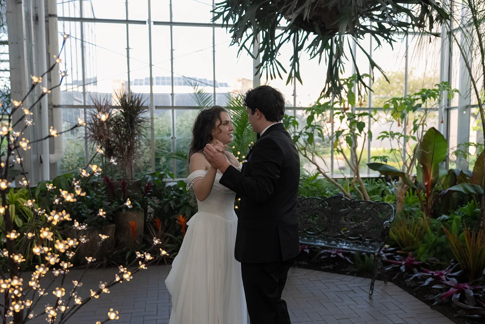 A bride and groom dancing together in a greenhouse or conservatory filled with plants, fairy lights, and natural light.