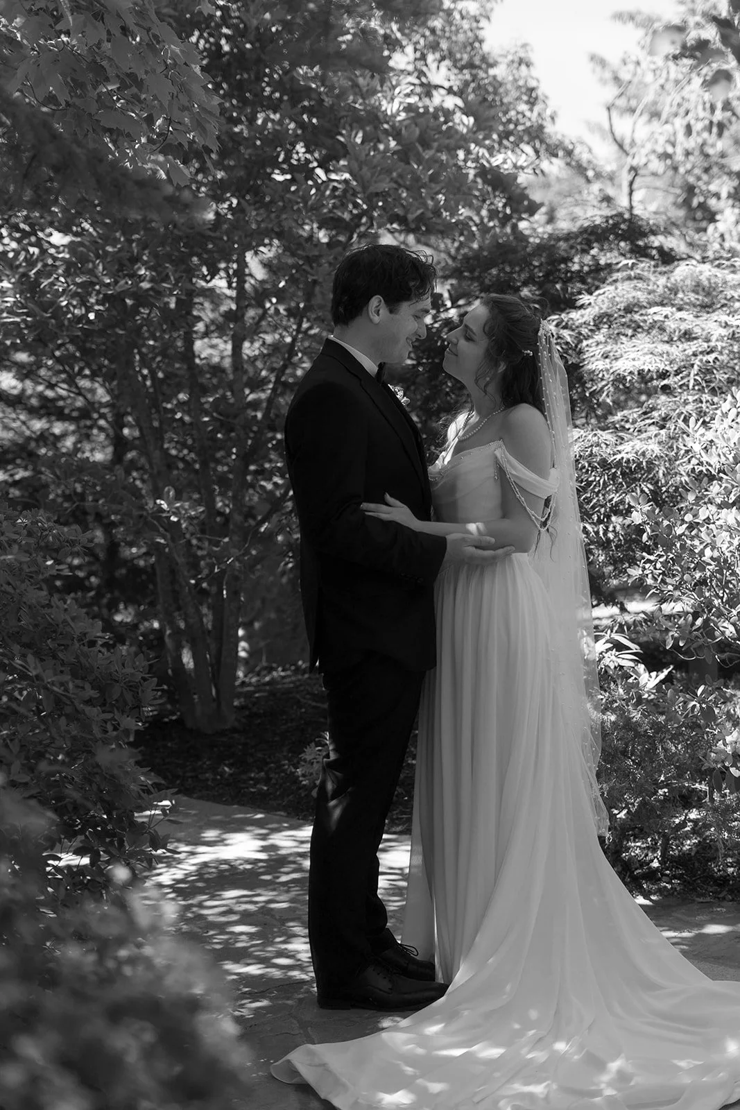 A black and white photo of a bride and groom standing close together outdoors, looking into each other's eyes, surrounded by trees and foliage.