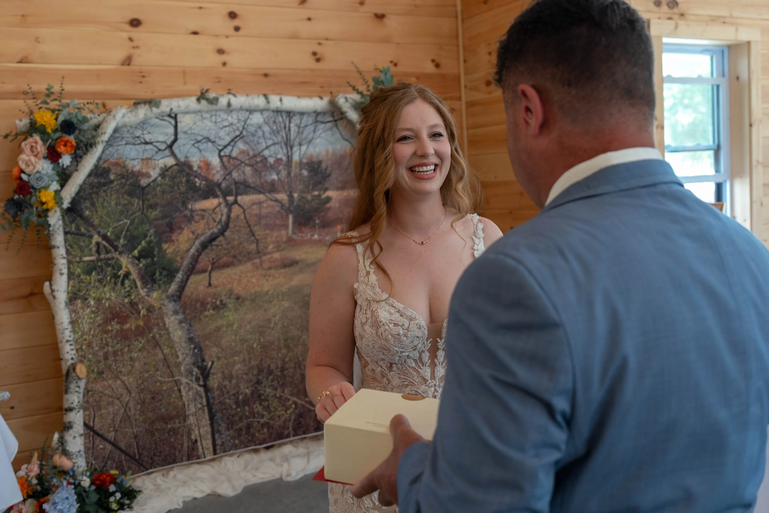 A bride and groom exchanging wedding vows indoors with a wooden-paneled wall and a scenic outdoor picture in the background. The bride wears a lace wedding dress and the groom wears a suit, and they are smiling at each other during the ceremony.