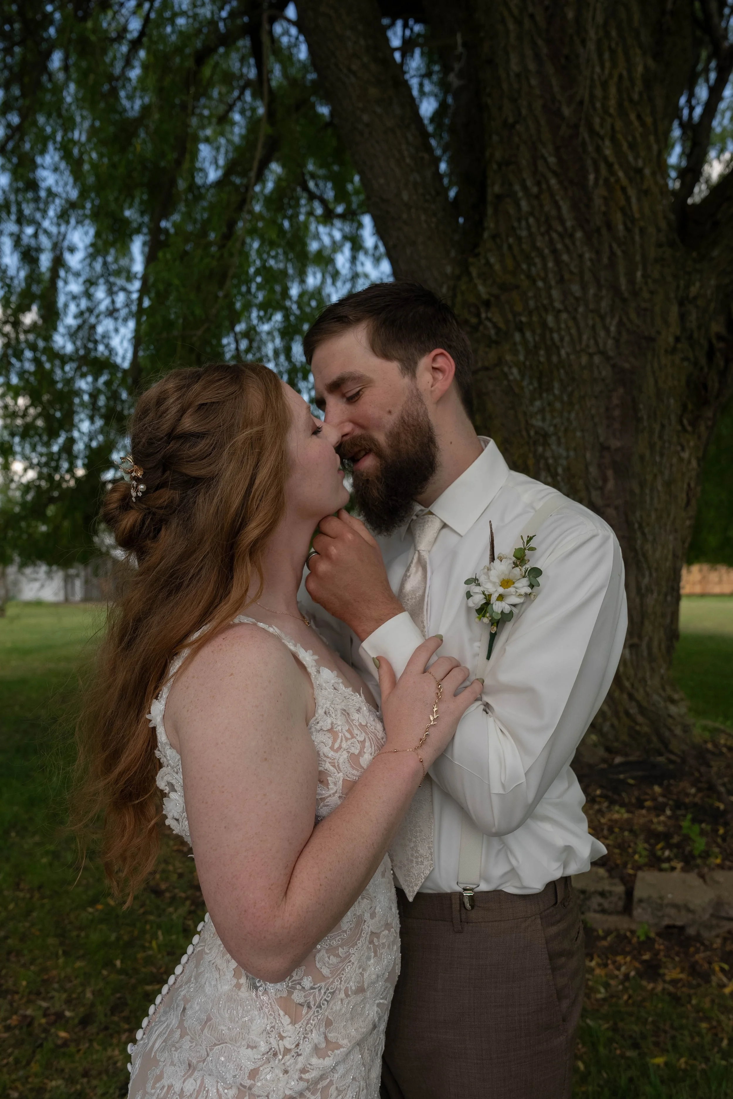 A couple sharing a close moment outdoors under a large tree, dressed in wedding attire; the woman has long red hair and a laced wedding dress, and the man has a beard, brown hair, a white shirt, and a boutonniere, with their faces close and eyes clos