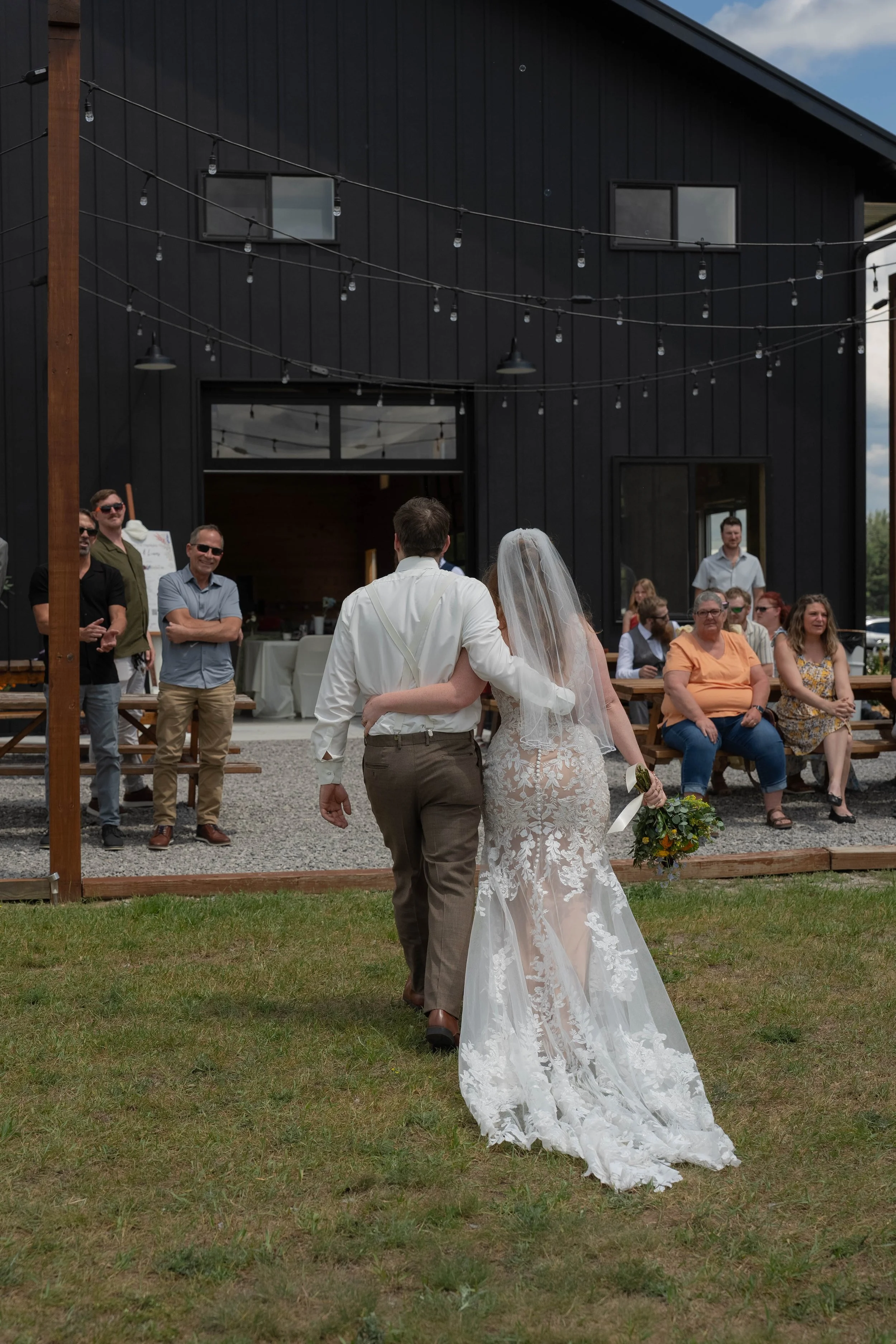 A bride and groom walking together at an outdoor wedding ceremony with guests seated on benches in the background, a modern black building in the distance, and string lights overhead.