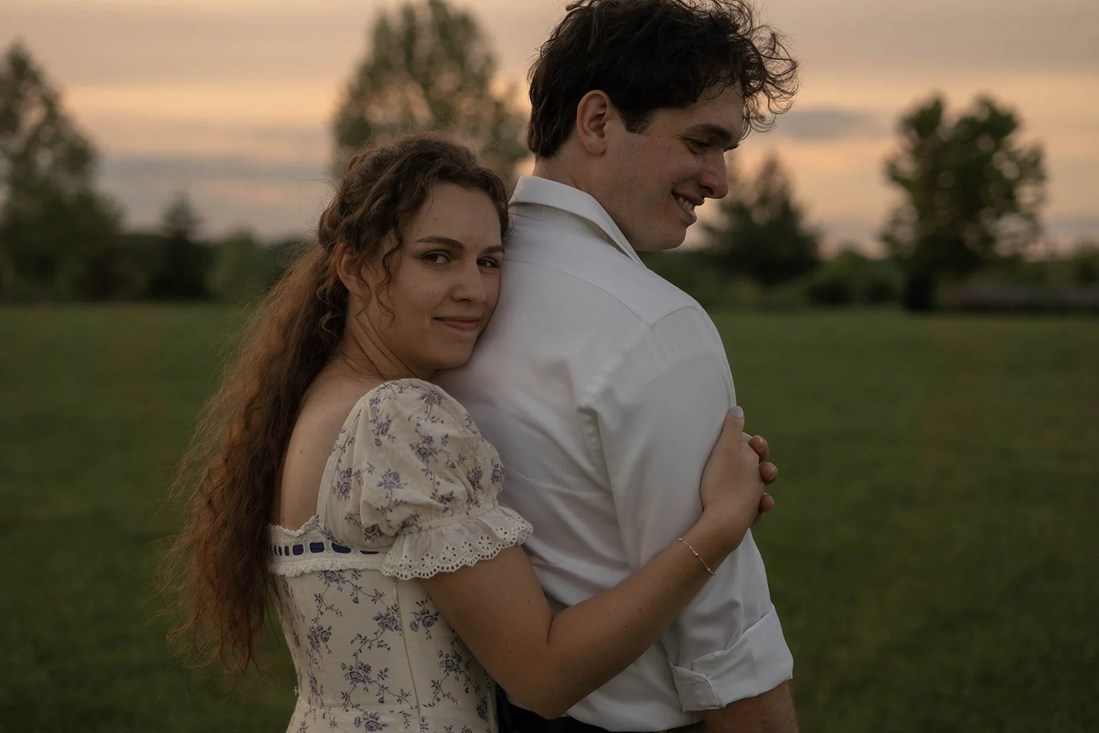A young woman with curly brown hair and a white floral dress embraces a young man with dark hair and a white shirt in a grassy field during sunset.