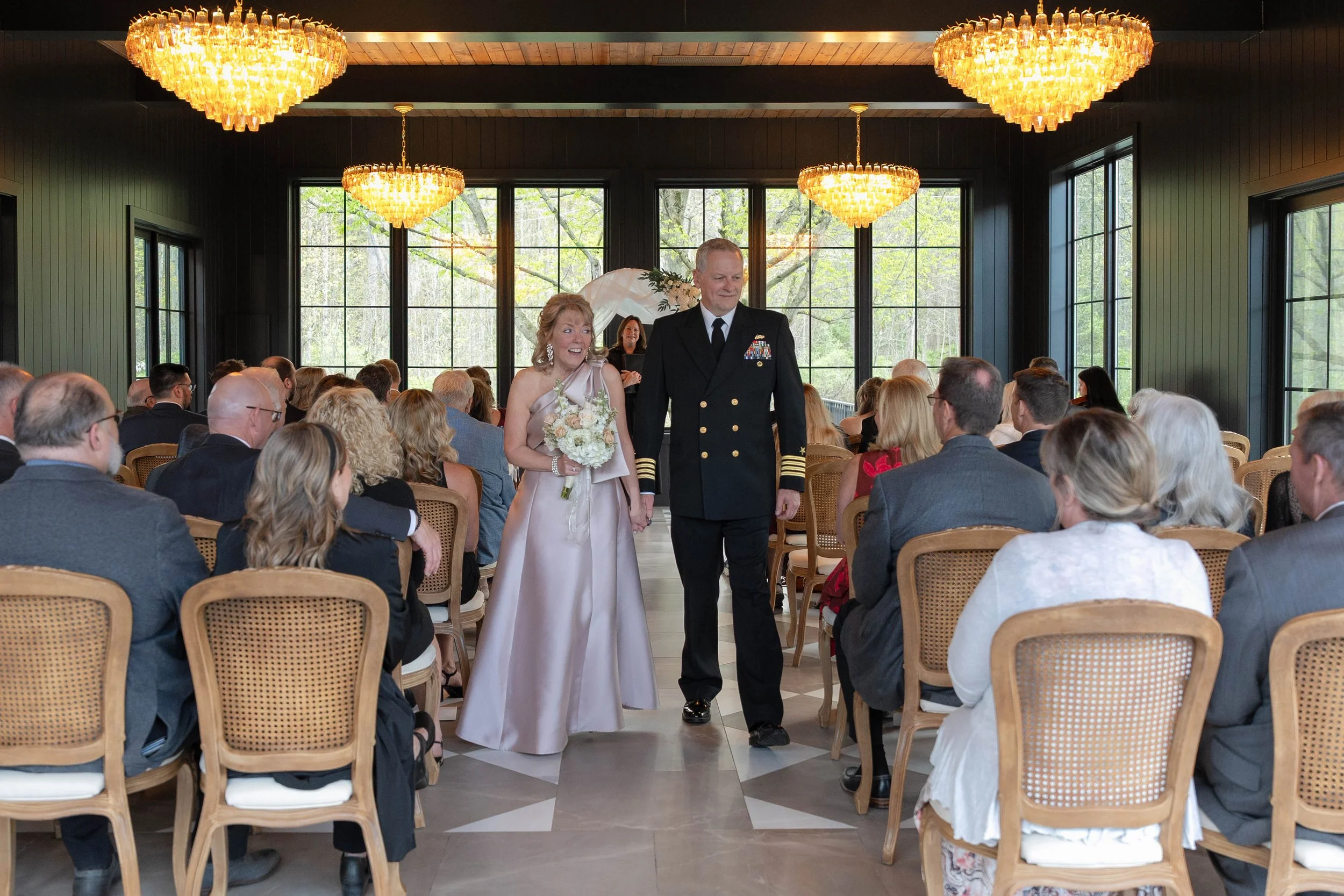 A bride and groom walking down the aisle of a wedding ceremony. The bride is in a satin gown holding a bouquet, and the groom is in a navy uniform. Guests are seated, and the setting features large windows and hanging chandeliers.