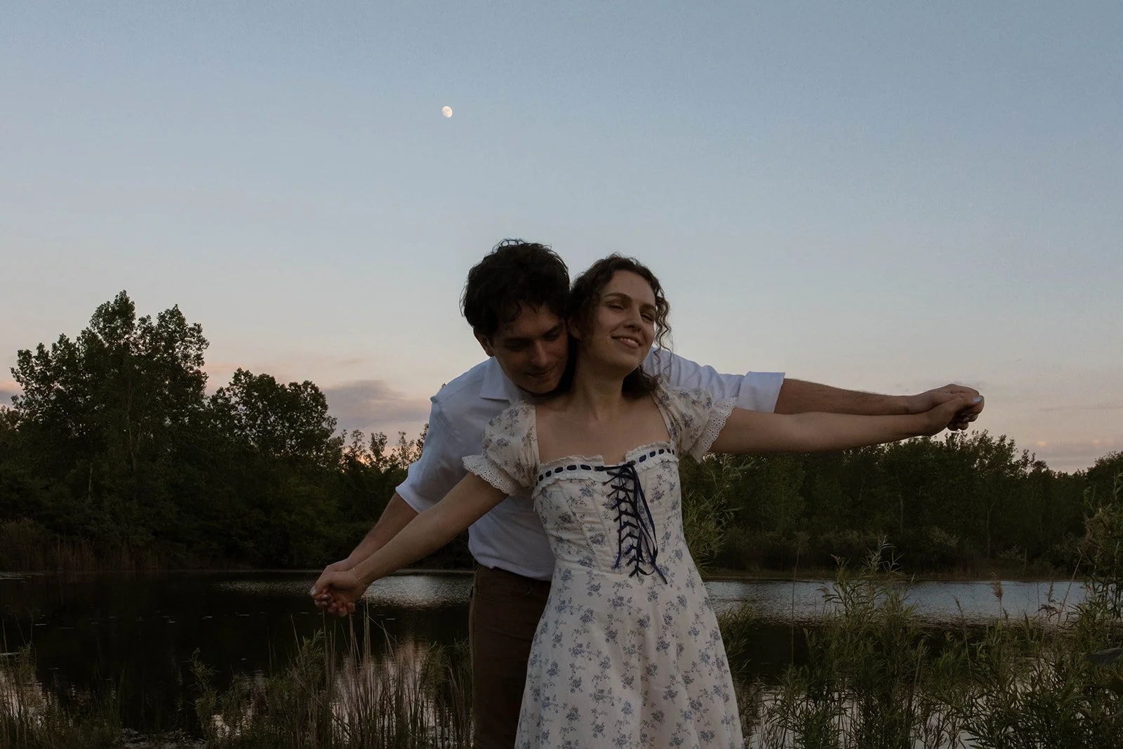 A young man and woman with arms outstretched by a river during sunset, with trees and the moon in the sky.