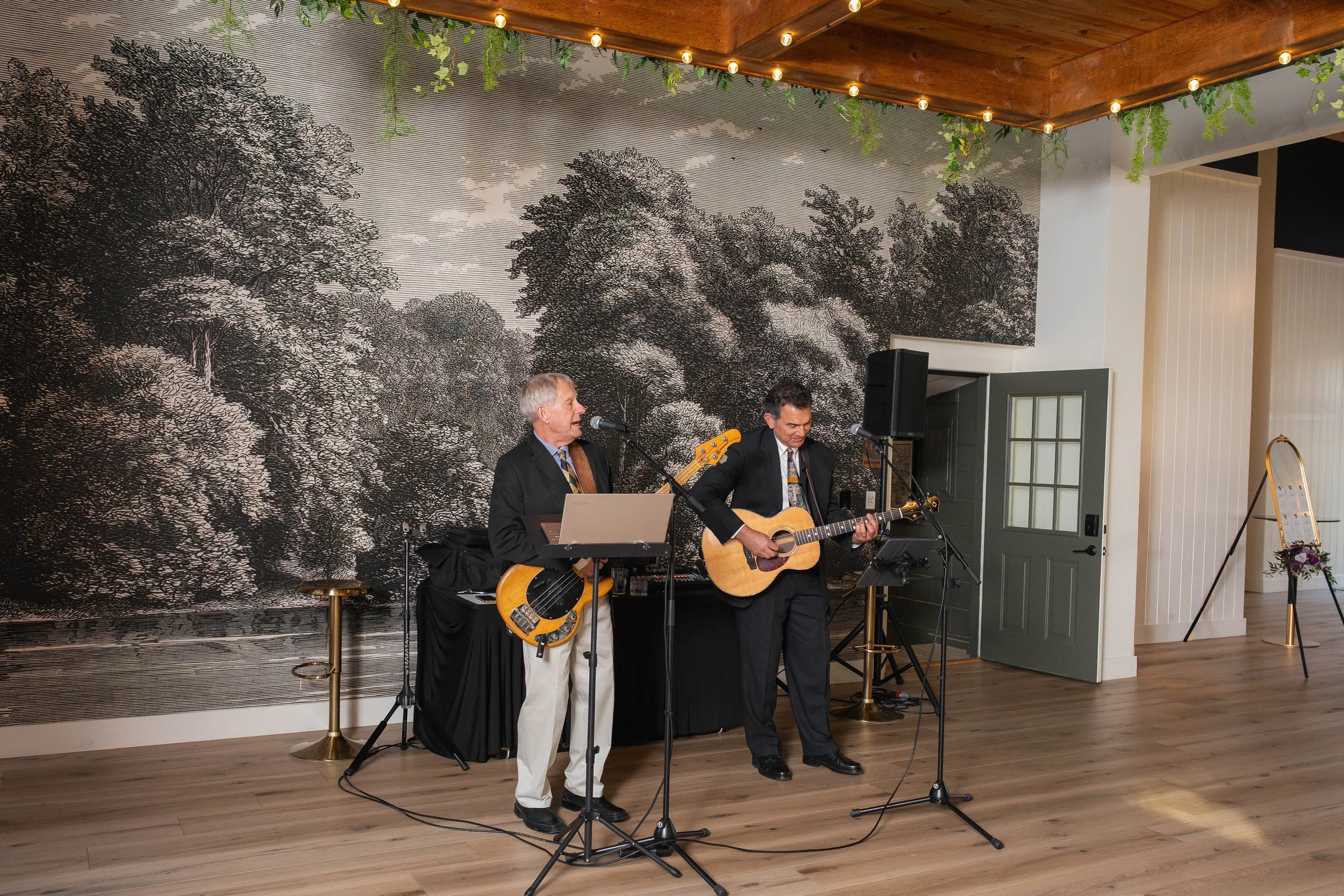 Two musicians in black suits playing guitars and singing in front of a large black and white tree mural, with a wood ceiling decorated with string lights and hanging green plants.