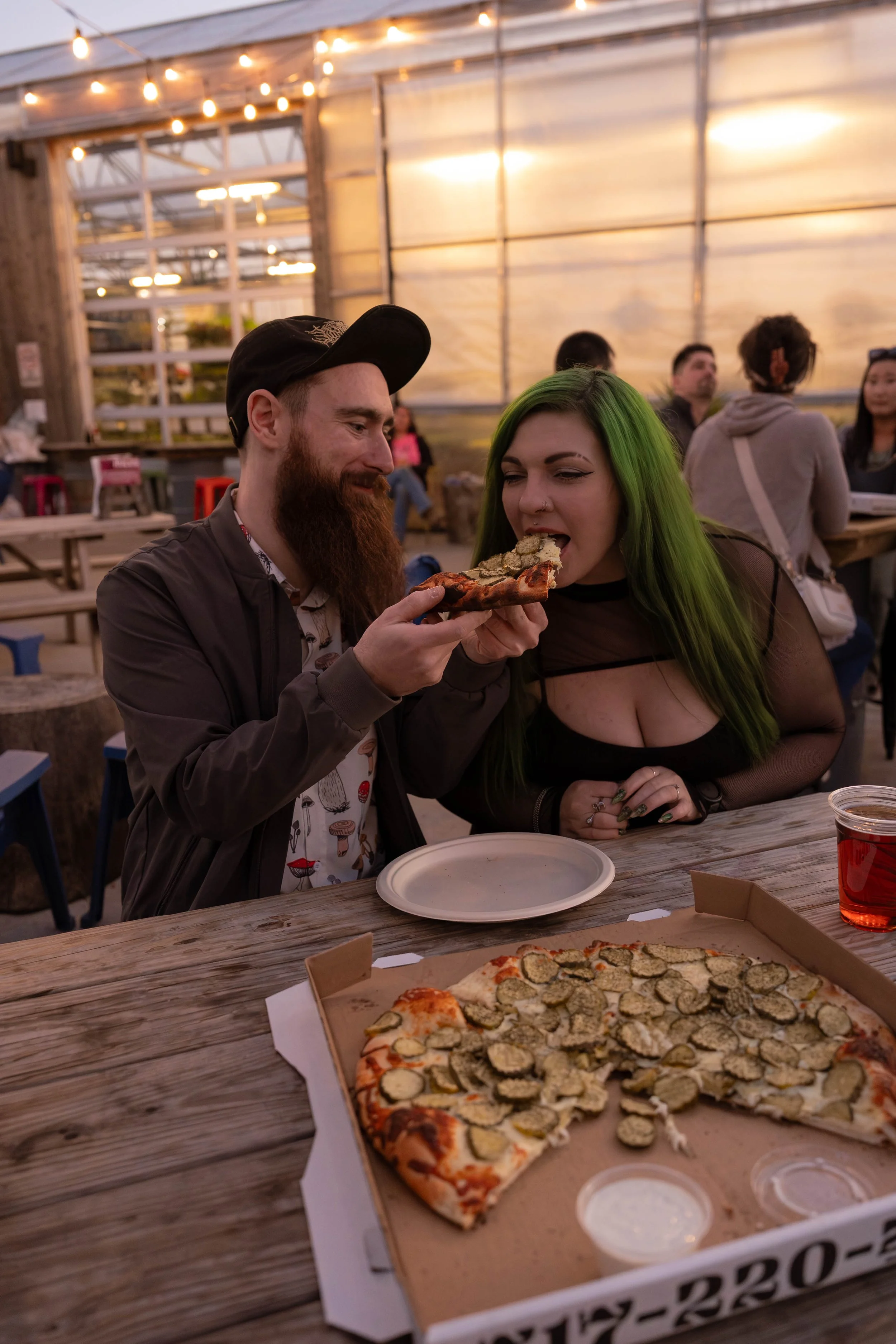 A man and woman sharing a slice of pizza at an outdoor dining area during the evening, with a pizza box and pizza on the table in the foreground.