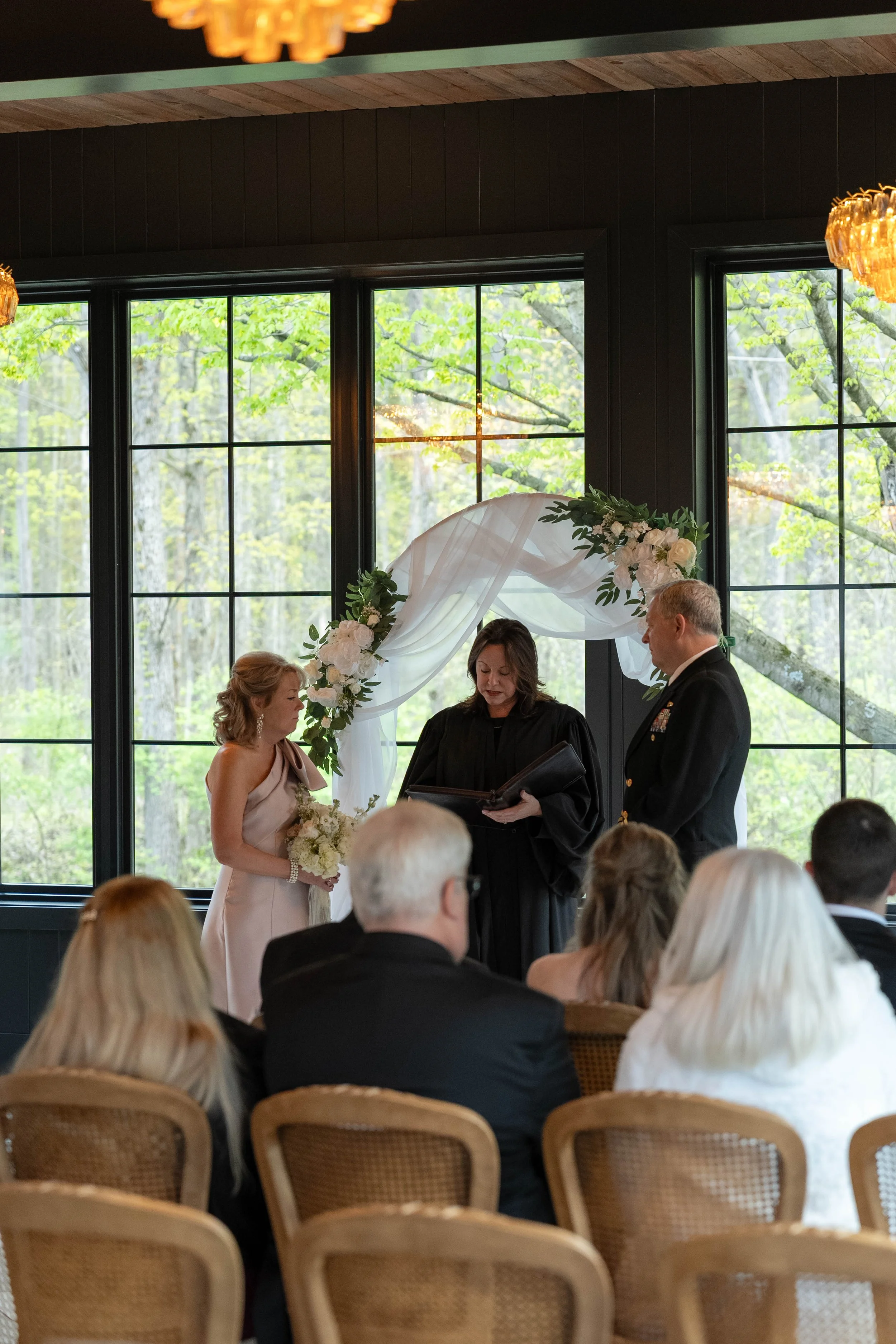 A wedding ceremony inside a building with large windows and natural light. A bride and groom stand in front of an officiant, with guests seated and watching.
