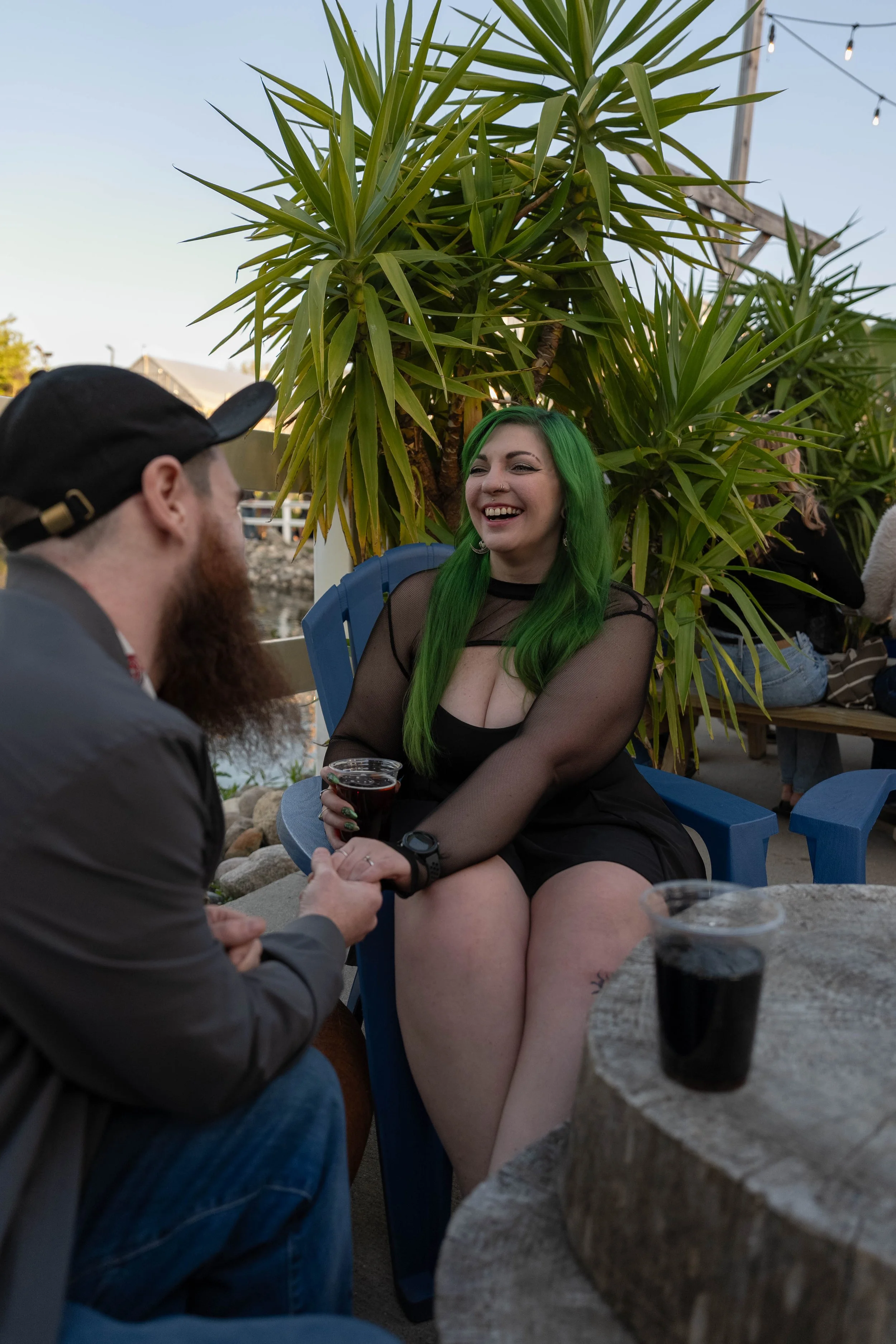A woman with bright green hair smiling and holding a drink, sitting on a blue chair outdoors at a social gathering, engaging in conversation with a bearded man wearing a black cap, with palm trees and string lights in the background.