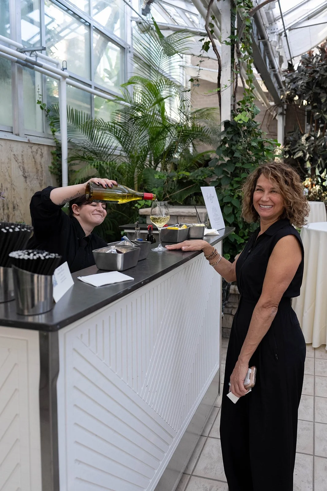 A woman in a black dress is smiling as she receives a glass of white wine from a bartender pouring wine from a bottle. The scene is set in a bright indoor space with green plants and glass walls.