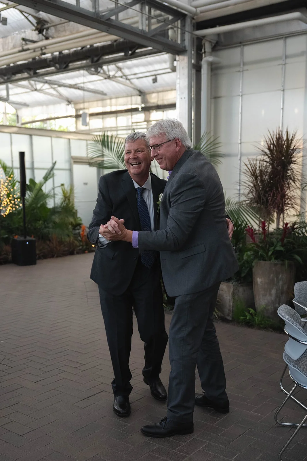 Two men in suits smiling and holding hands in a dance or greeting, indoors with plants and decorative lights.