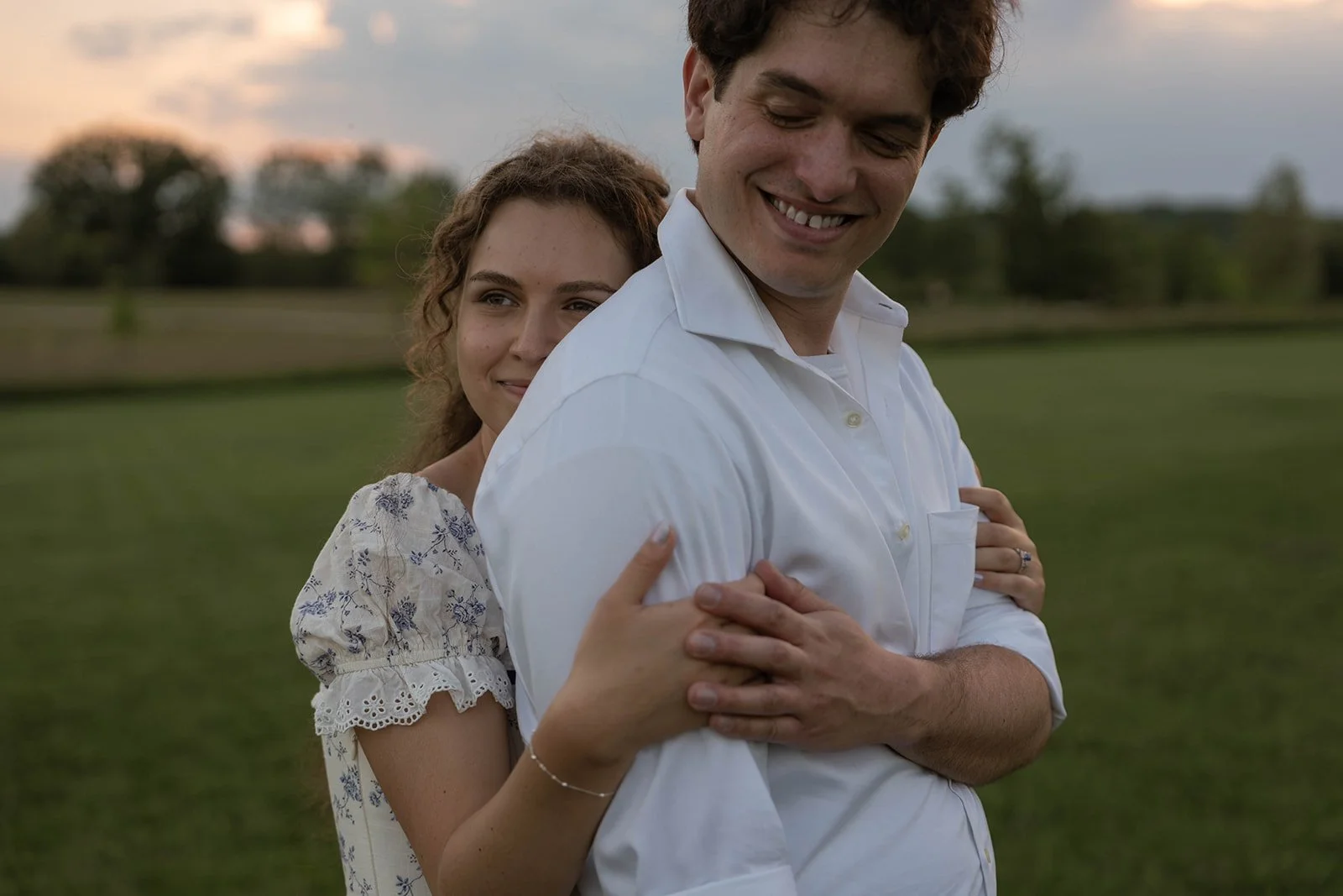 A young woman with curly hair and a white floral dress hugging a smiling young man with dark curly hair in a white shirt, standing outdoors on a grassy field during sunset.