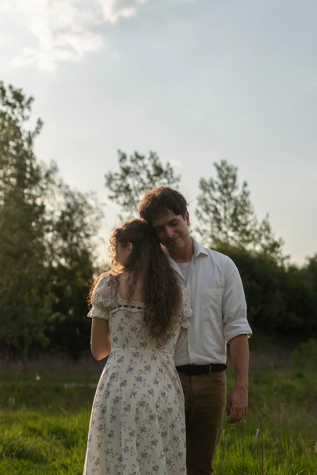 A young couple holding each other in a grassy field during sunset, with trees and a partly cloudy sky in the background.