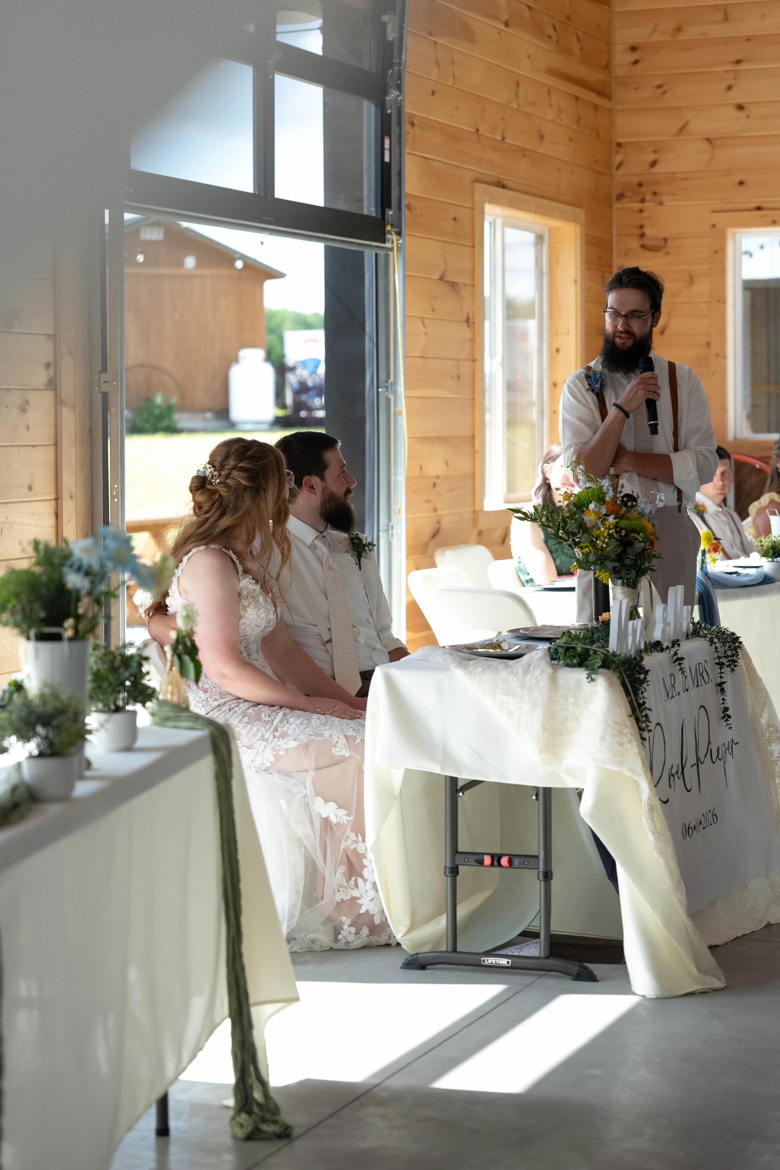 A wedding reception inside a wooden building with natural light. A man with a beard, glasses, and suspenders is giving a speech into a microphone next to a decorated table that has a sign reading 'MR. & MRS.' and a bouquet of flowers. The bride and g