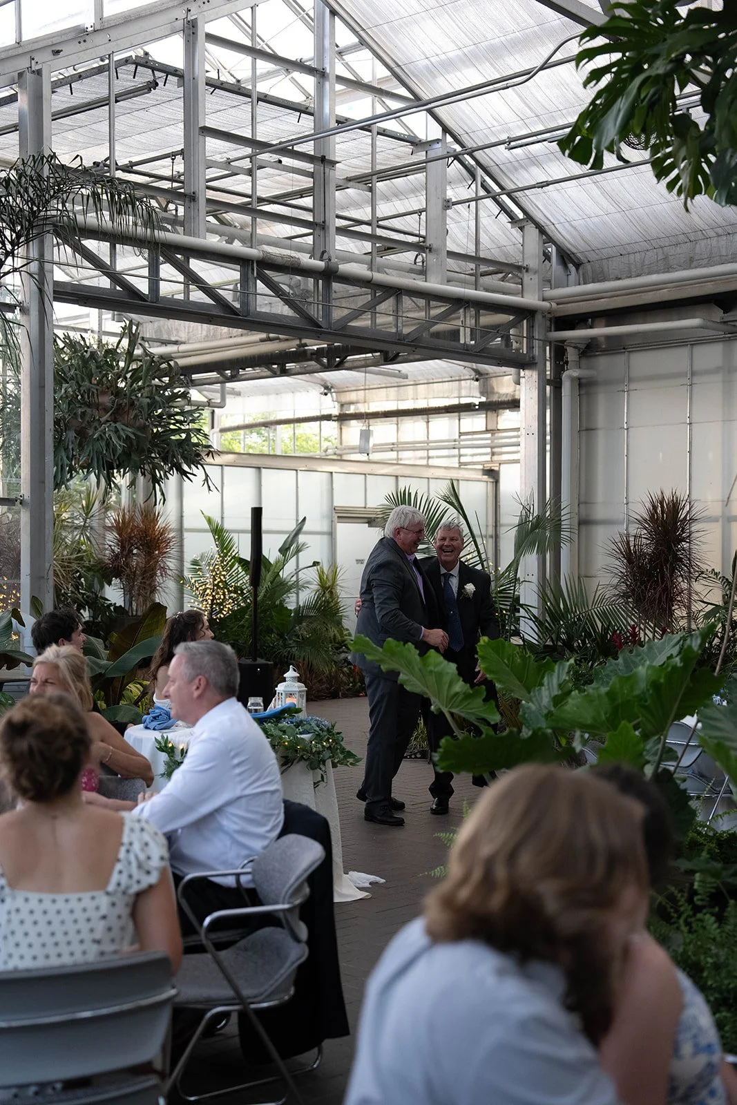 Two men in suits are smiling and holding hands in a greenhouse decorated with lush green plants, with seated guests at tables around them, suggesting a wedding or celebration.