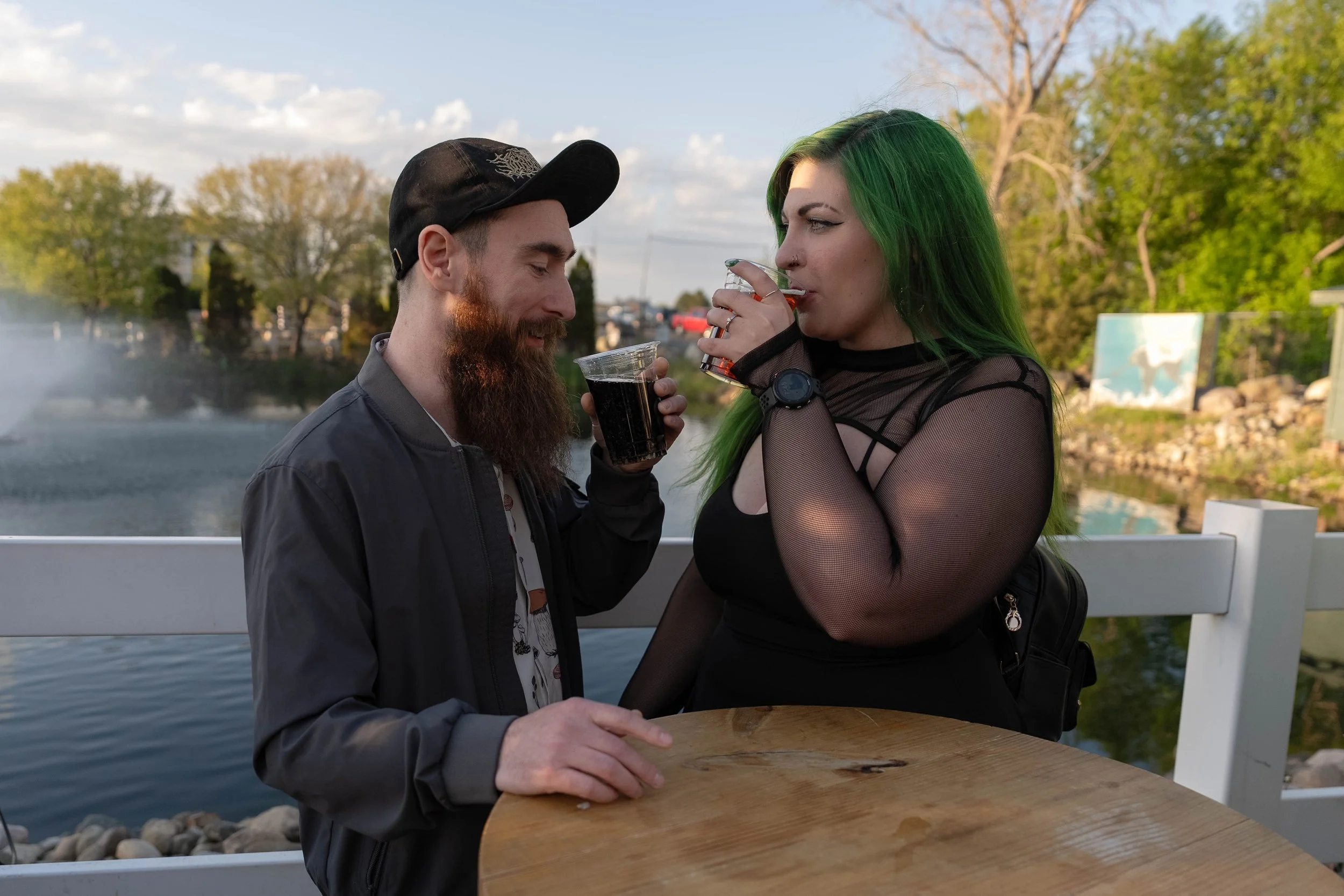 A man with a beard and a black cap is holding a drink, and a woman with green hair is sipping from a clear glass while standing outside near a body of water with trees and a bridge in the background.