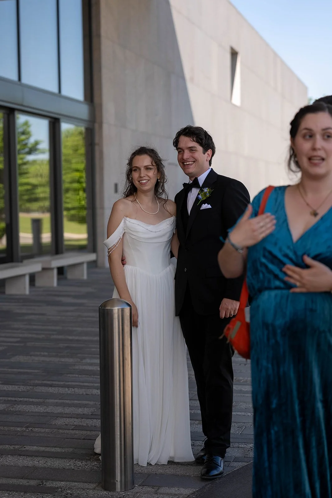 A bride and groom smiling together outdoors near a modern building, with a woman in a blue dress and a red bag nearby.